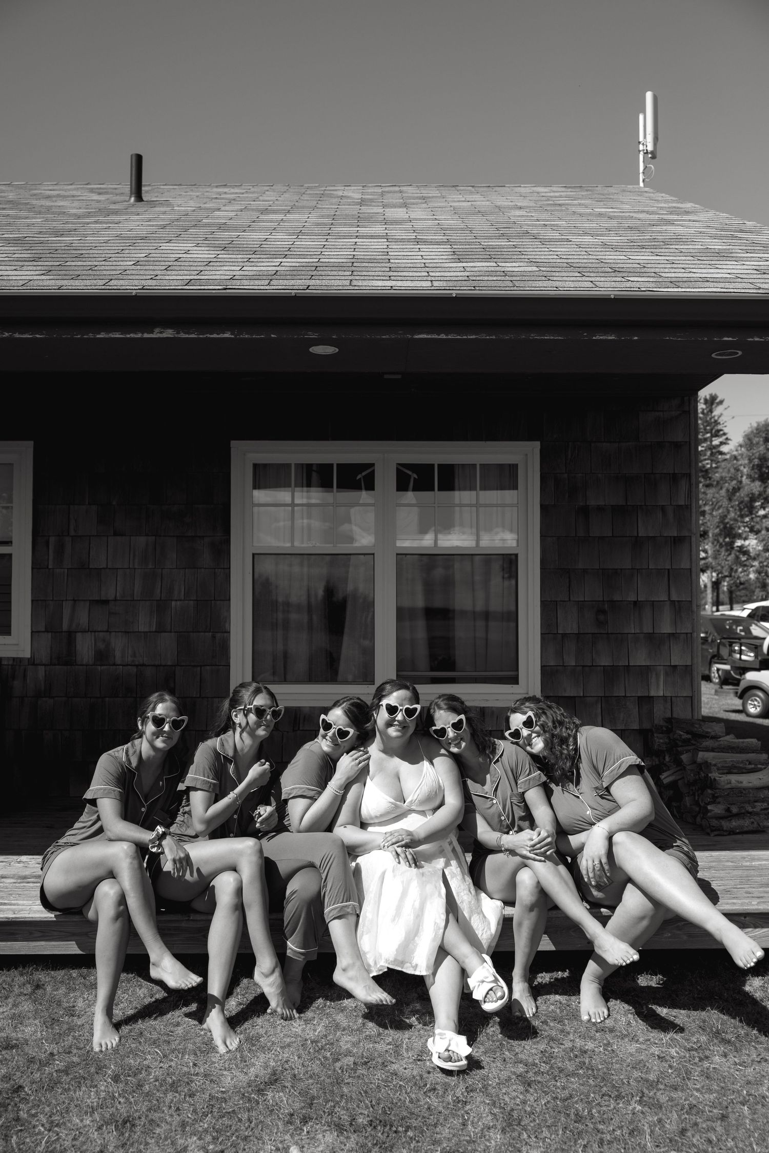 Wedding party sits together on cabin steps in black and white photo, laughing and wearing sunglasses.