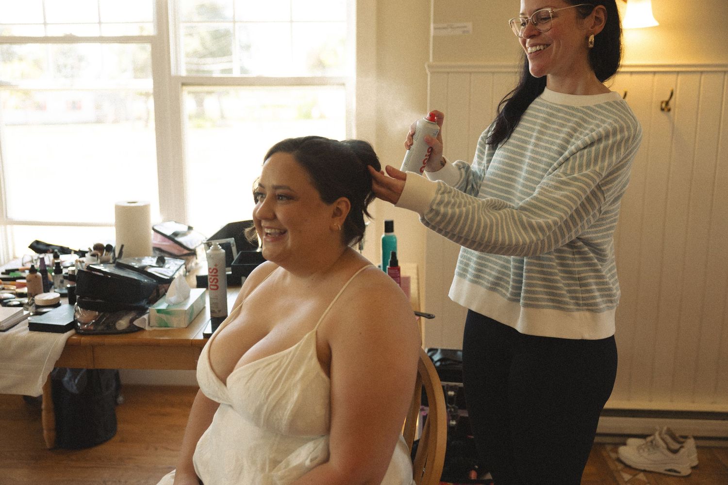 A bride smiling as a stylist applies hairspray during wedding preparations indoors.
