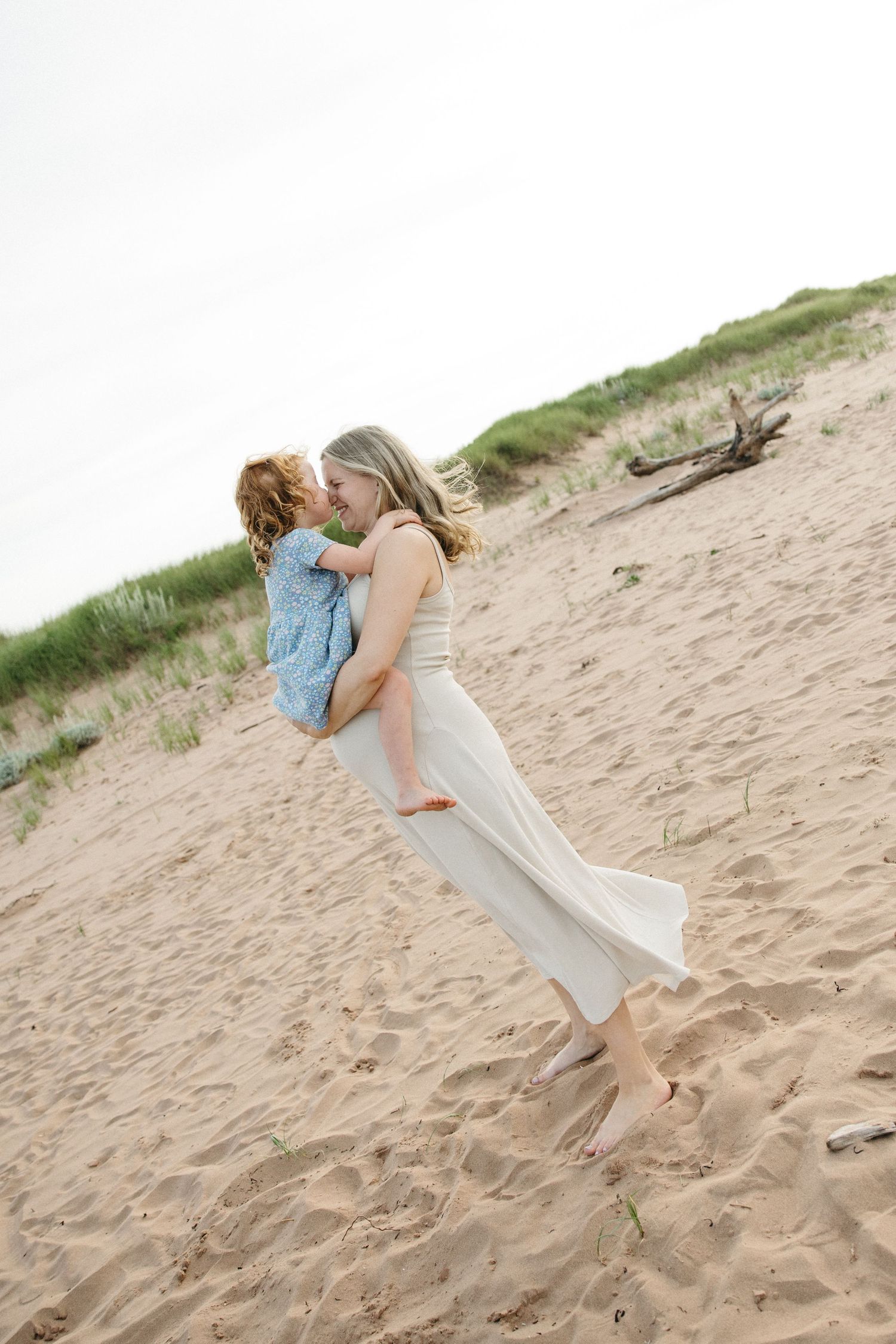 A joyful mother in a white dress lifts and embraces her young child on a sandy beach.