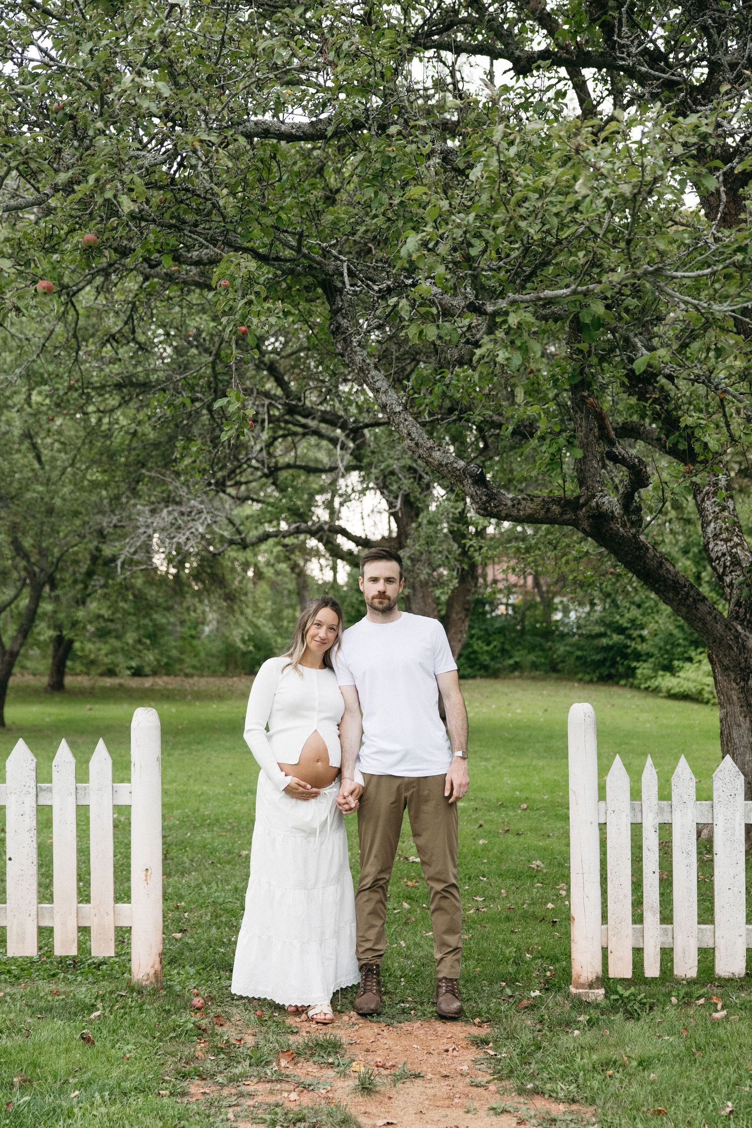 Pregnant couple standing together by white picket fence under a large garden tree.