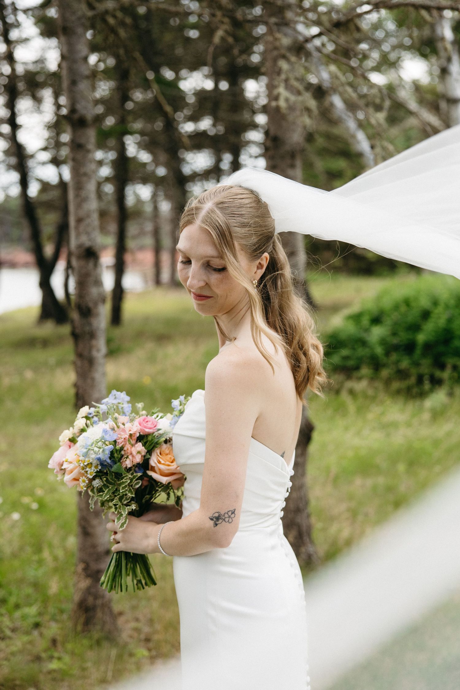 Bride in strapless white gown holding colorful bouquet with veil blowing in wind among pine trees.