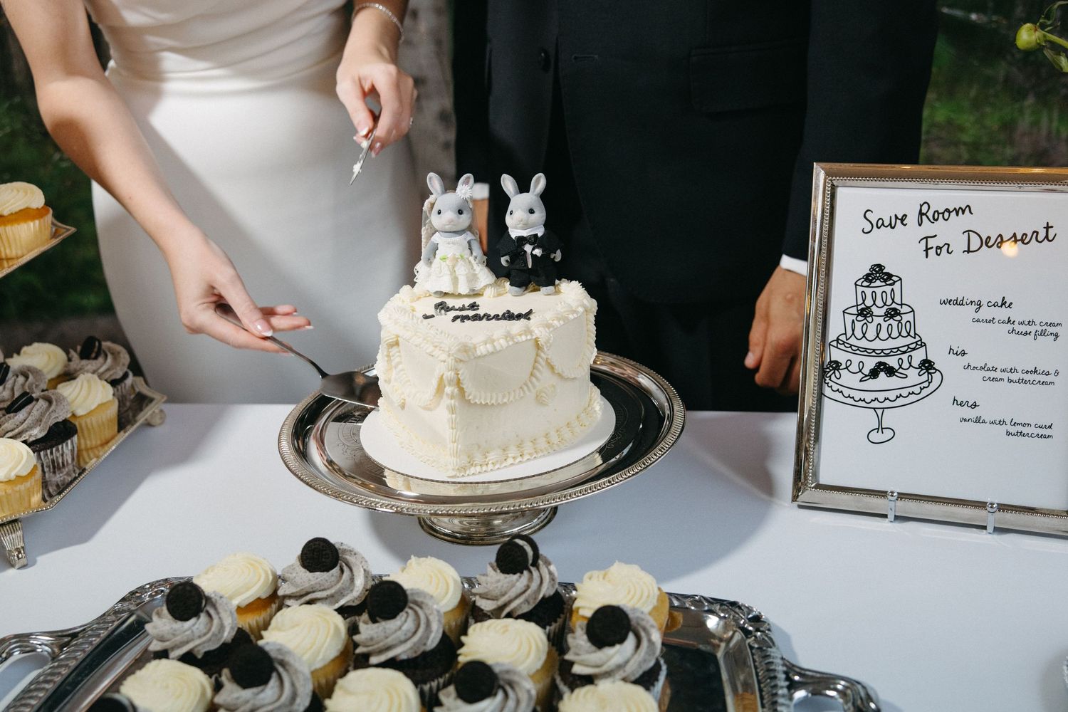 Bride and groom cut small white wedding cake with bunny toppers at elegant dessert table.