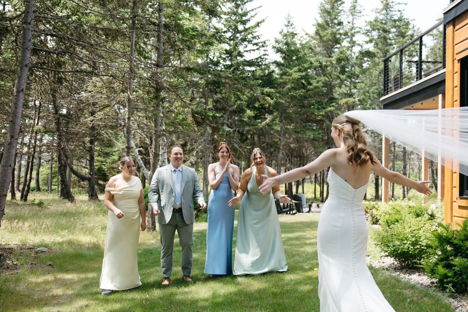 Bride in white gown with veil reveals herself to bridesmaids outdoors near pine trees.