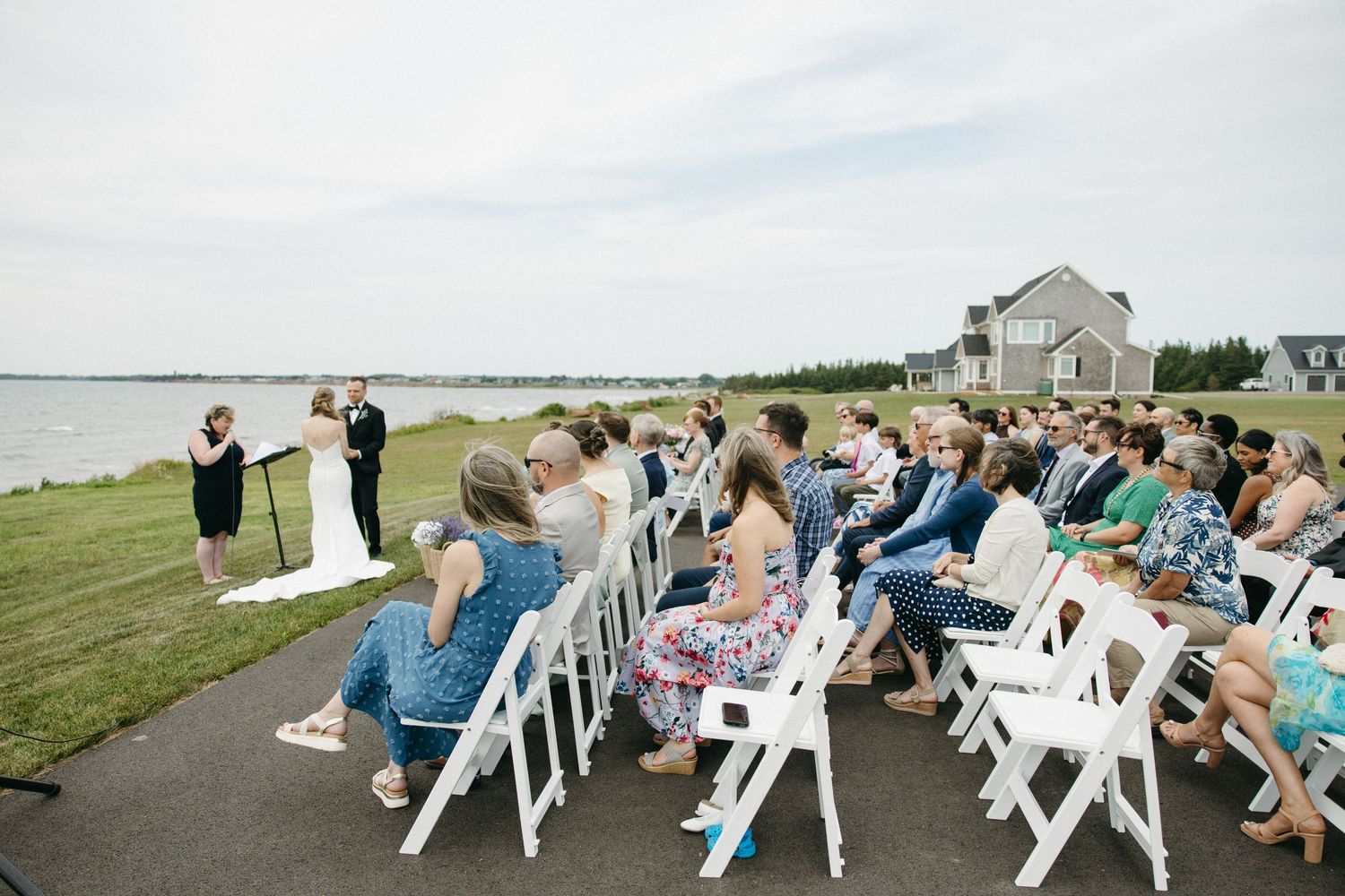 Outdoor waterfront wedding ceremony with guests seated in white chairs facing officiant and couple.