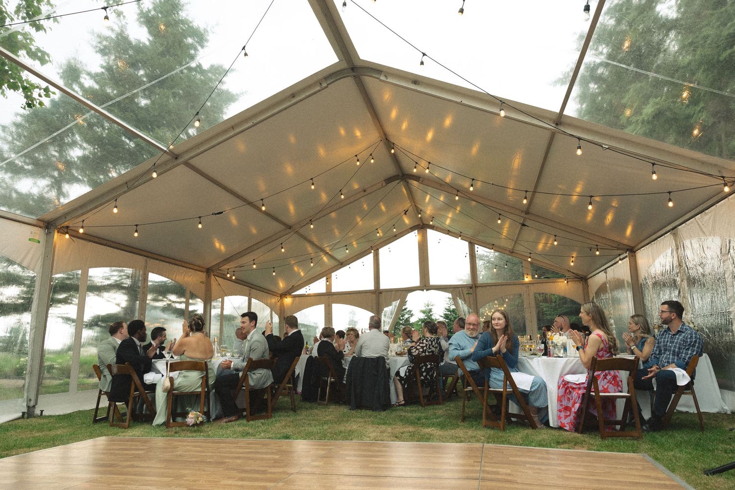 Wedding reception guests dining under a clear tent with string lights on a wooden dance floor outdoors.