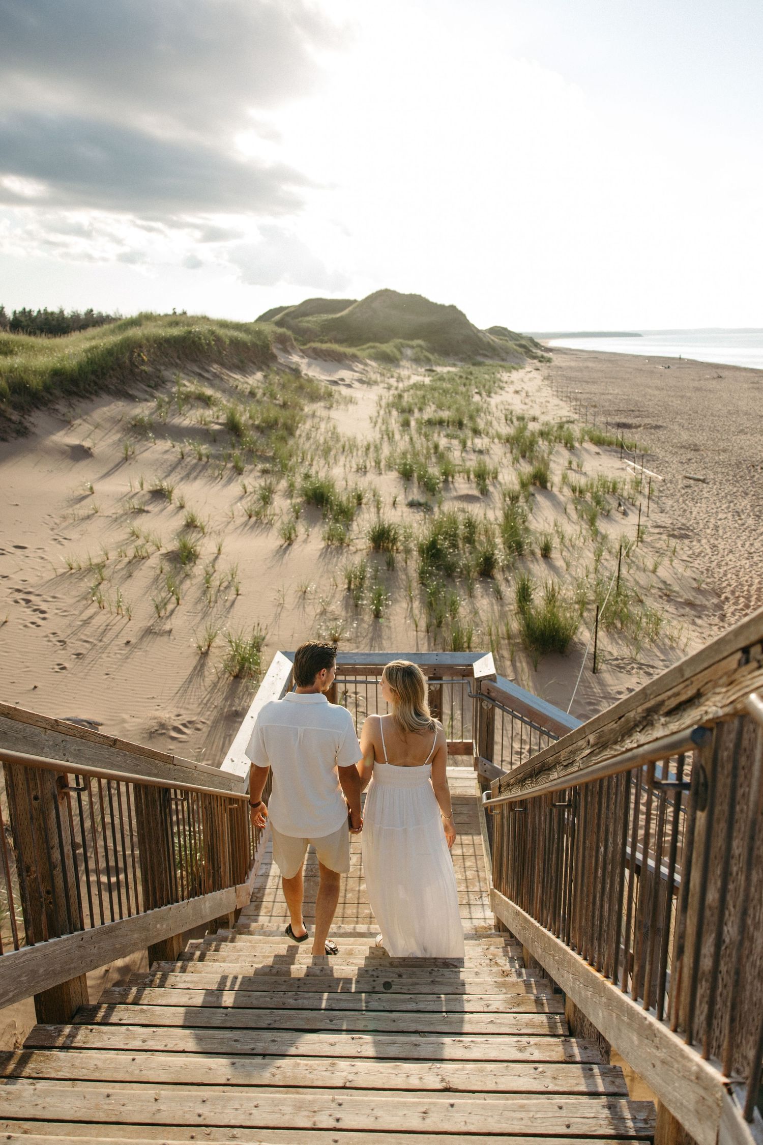 Couple holding hands walking down wooden beach stairs toward sandy dunes on a cloudy day.