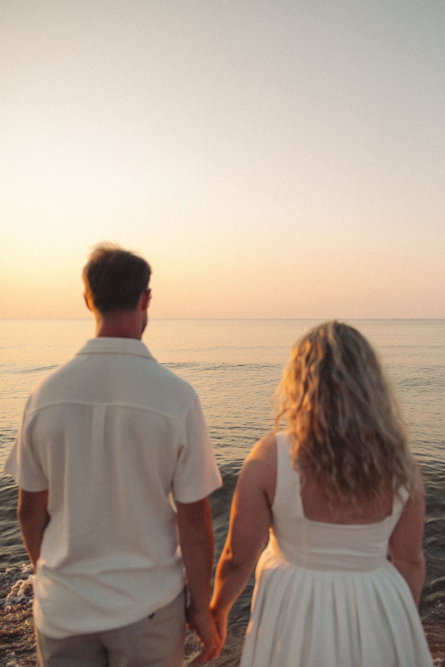 Couple in white outfits holding hands facing the ocean at a warm golden sunset.