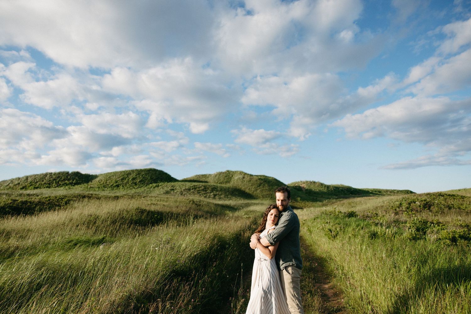 Couple embracing on a scenic green hillside path under a beautiful cloudy blue sky.
