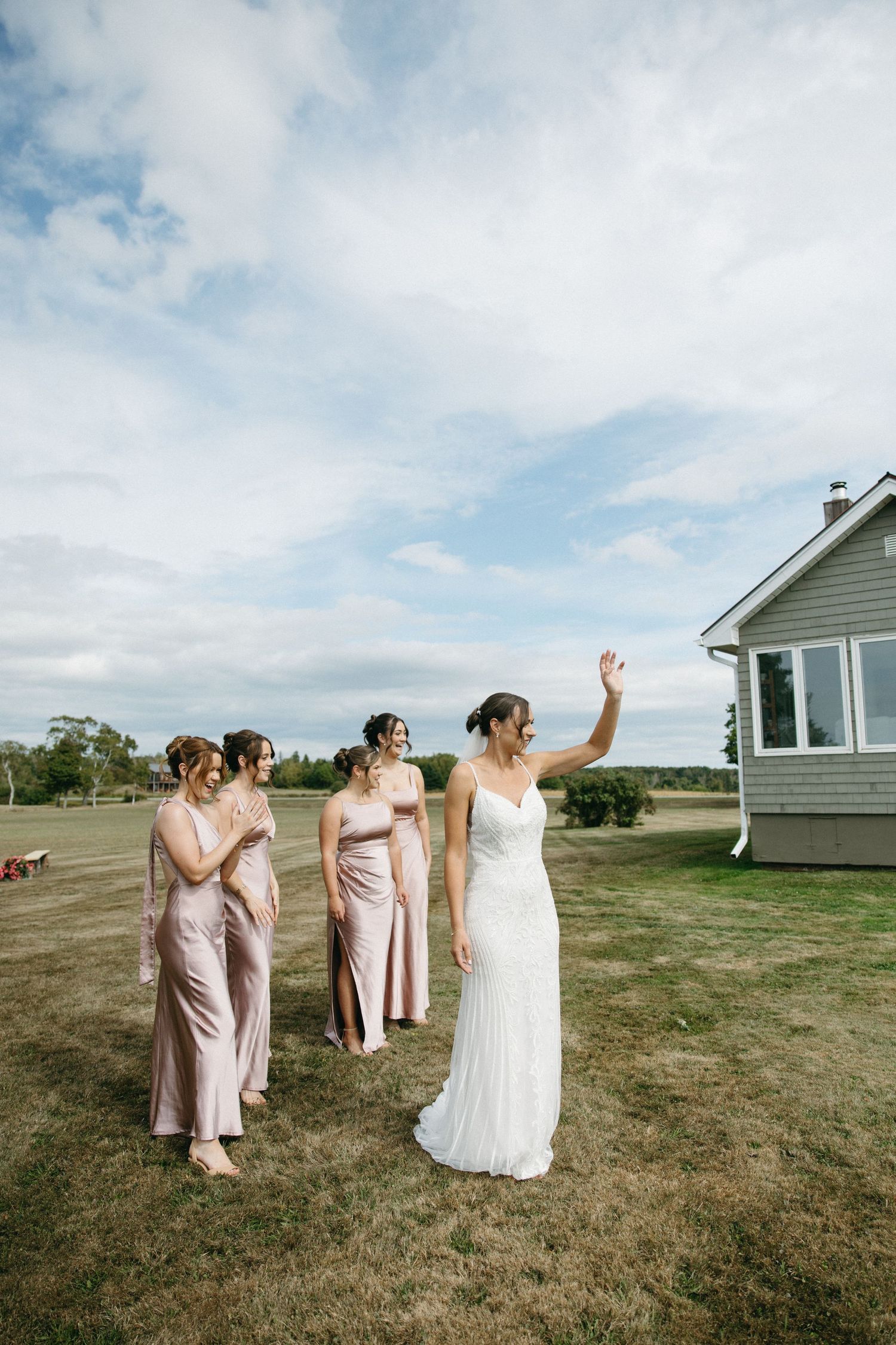 Wedding party in pink dresses and white gown posing on a grassy hill with houses in background.