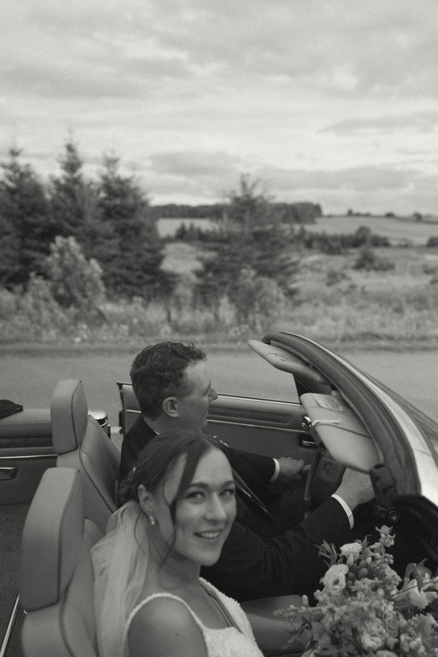 Wedding couple smiles while sitting in a convertible car with floral bouquet on a cloudy day.