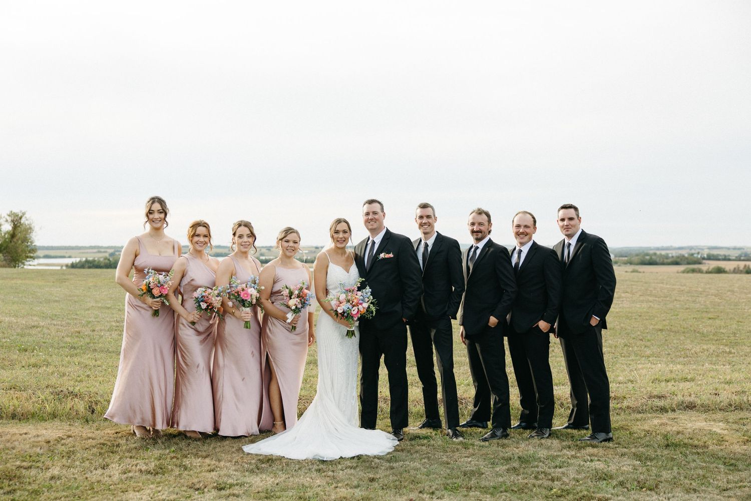 Wedding party in blush pink dresses and black suits poses together in an open field.