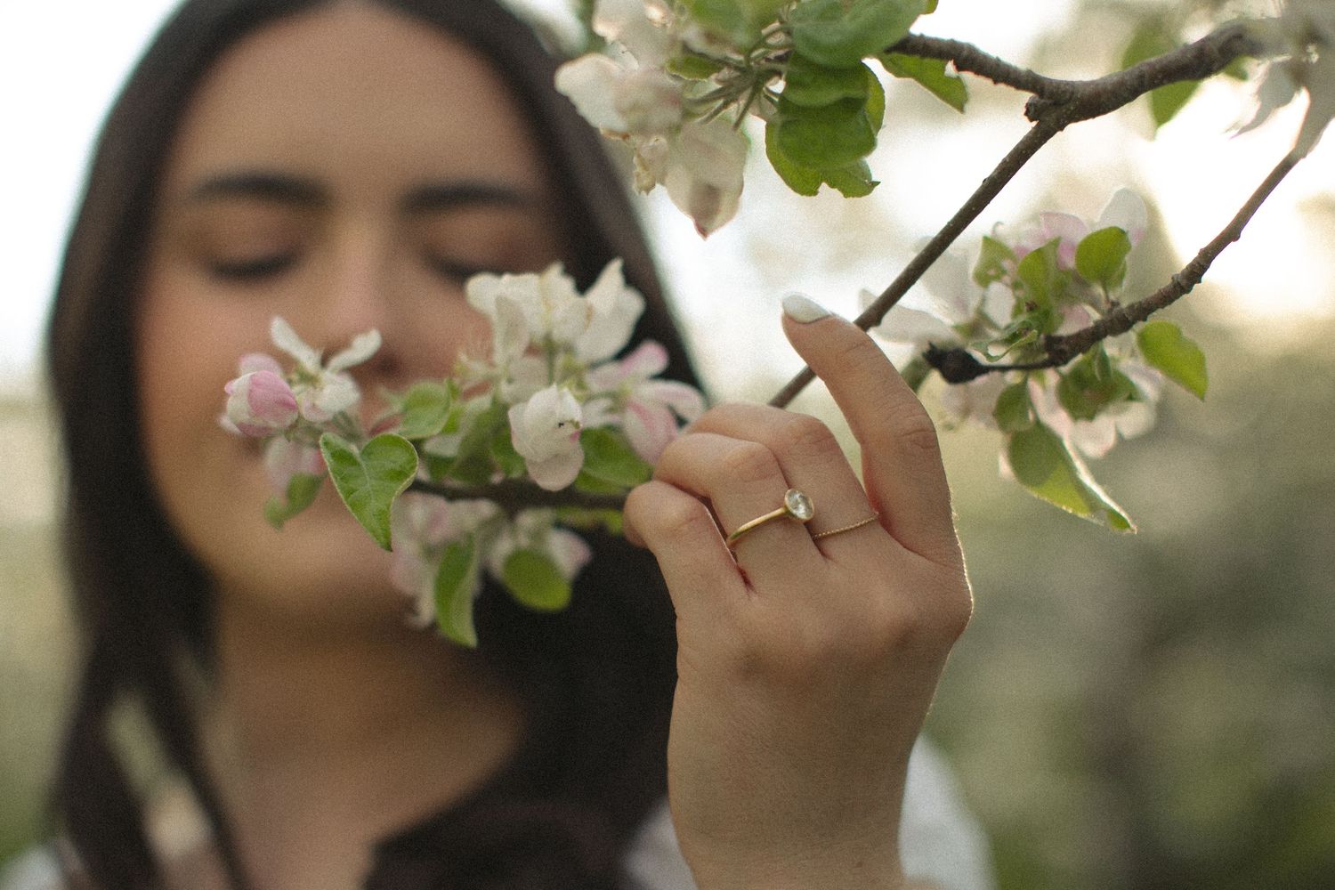 Woman with closed eyes smelling white spring blossoms on a tree branch, wearing a delicate gold ring.