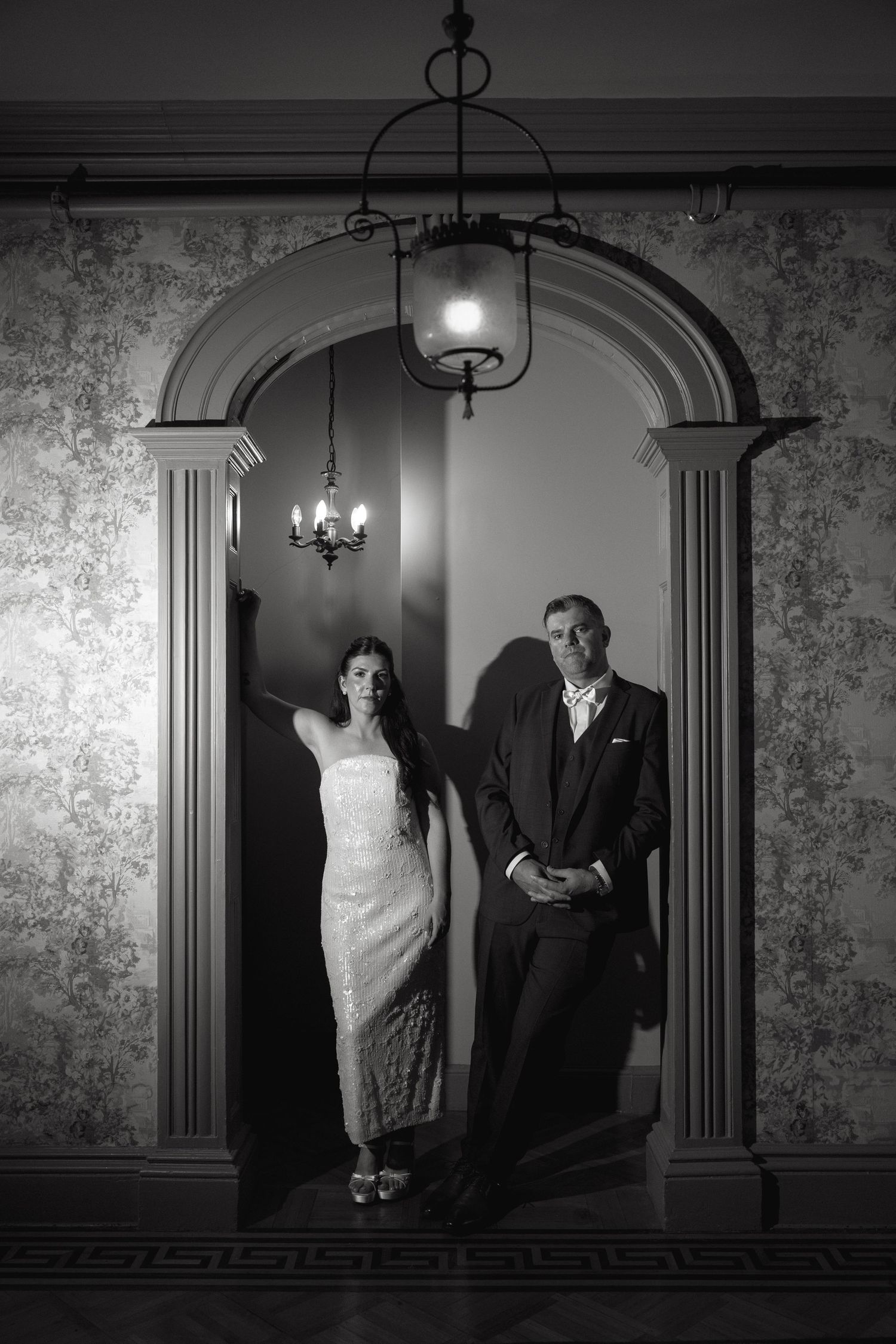 A black and white portrait of a bride and groom leaning against an ornate arched doorway beneath a vintage hanging lantern.