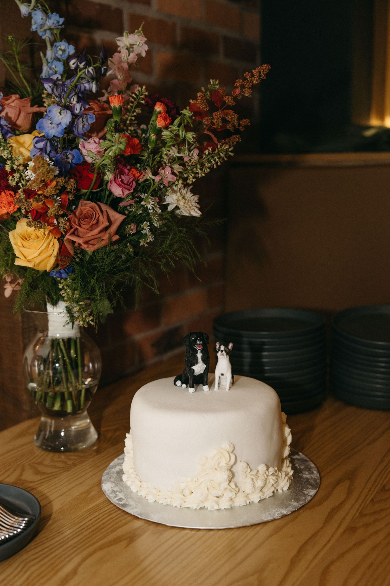 White fondant wedding cake with two dog figurine toppers on a wooden table beside a vase of colorful wildflowers and stacked dark plates.