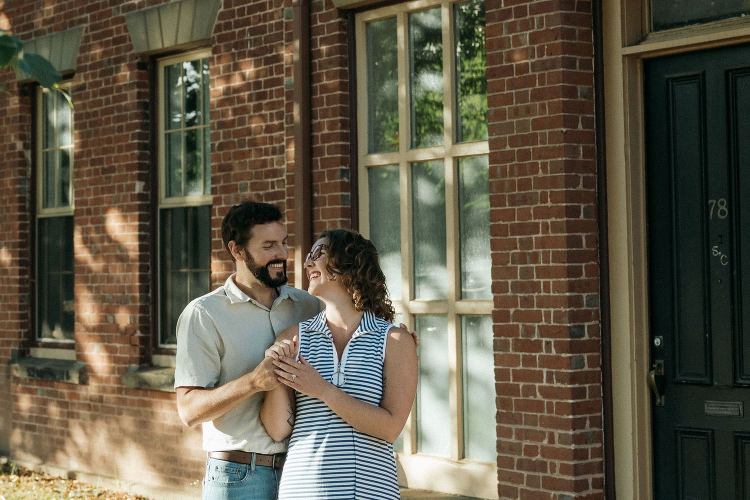 A couple embraces in front of a historic brick building with dappled sunlight.