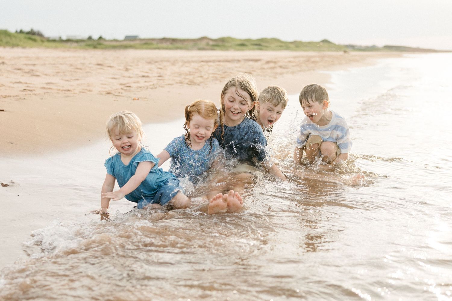 Five joyful children laugh and splash together in shallow ocean waves on a sandy beach on a cloudy day.