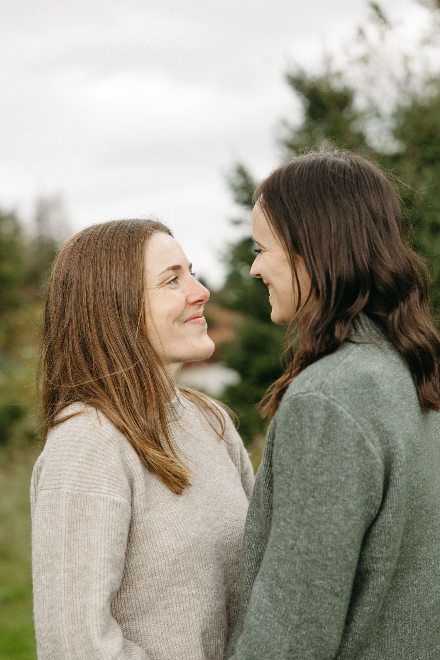Two smiling women in cozy sweaters stand close together facing each other outdoors during an autumn couples portrait session.
