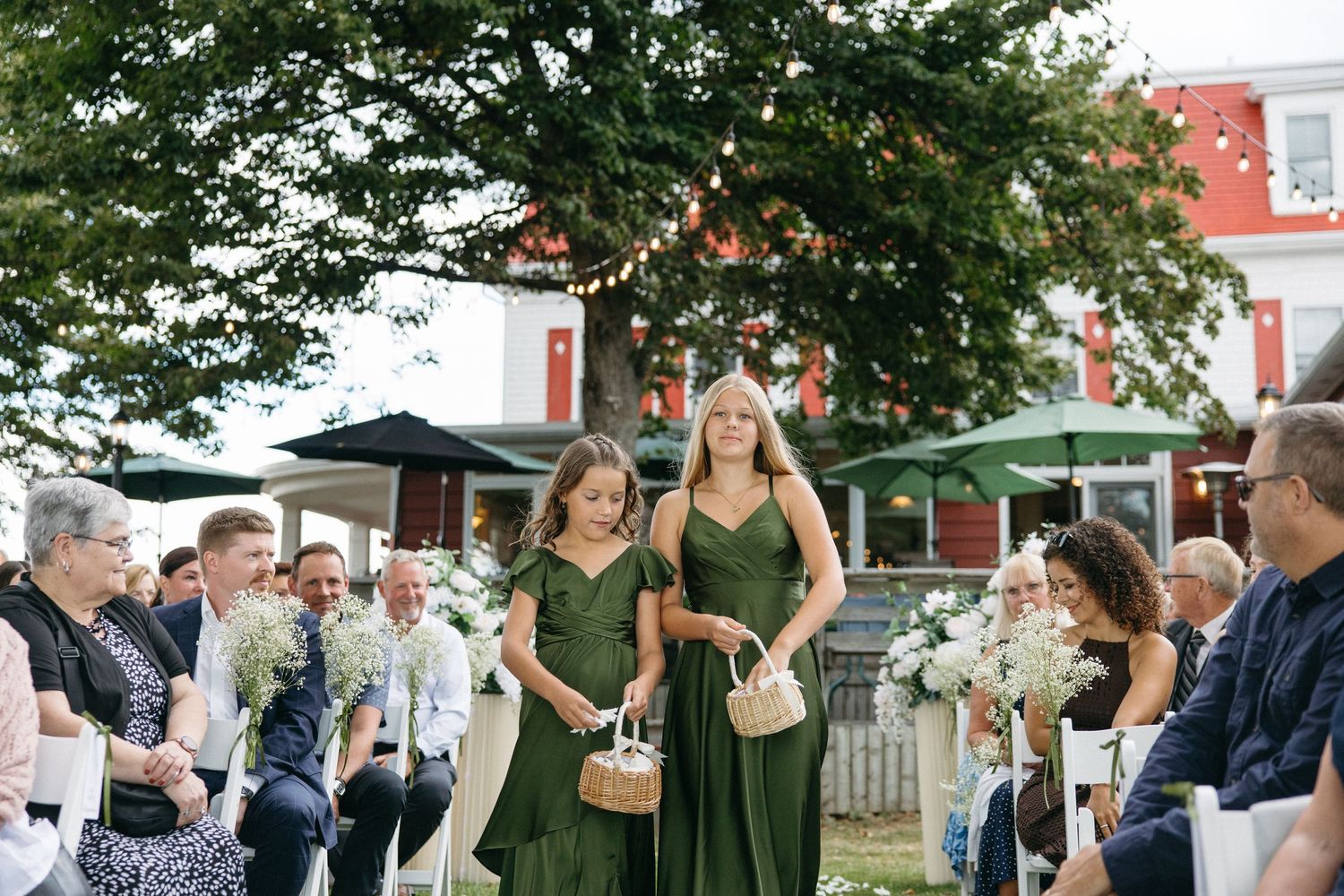Two flower girls in olive green dresses walk down an outdoor wedding aisle under a large tree.