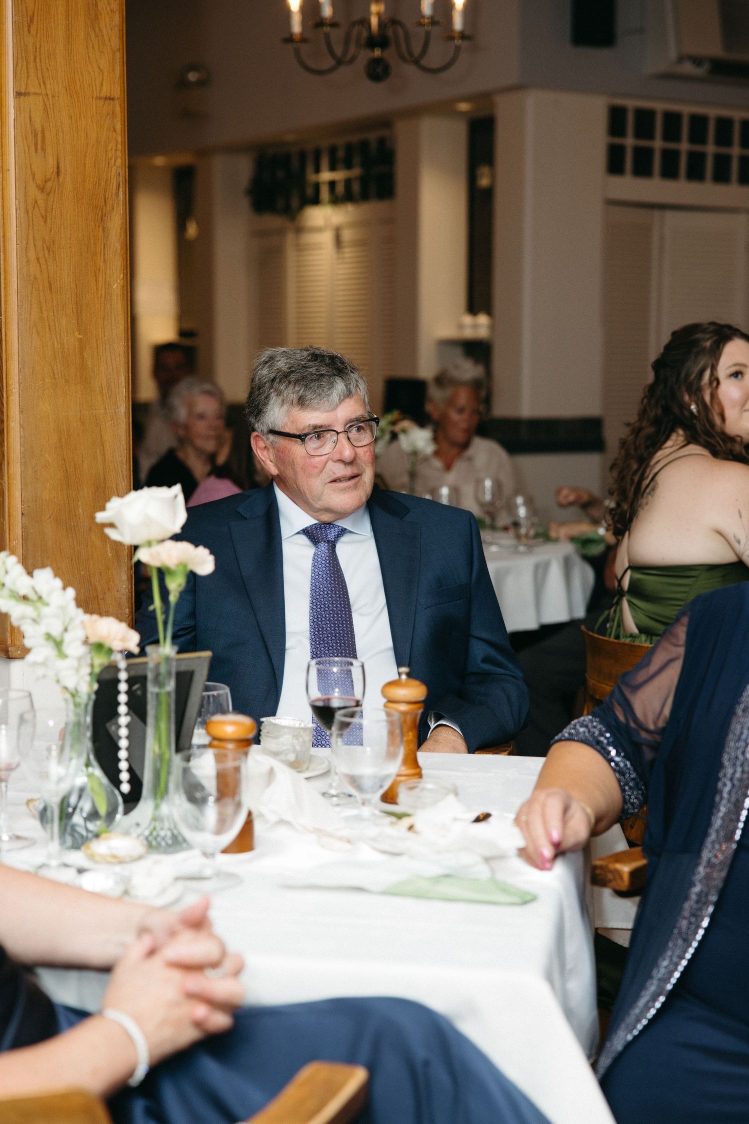Older gentleman in navy suit seated at elegantly set wedding reception table with white floral arrangements.