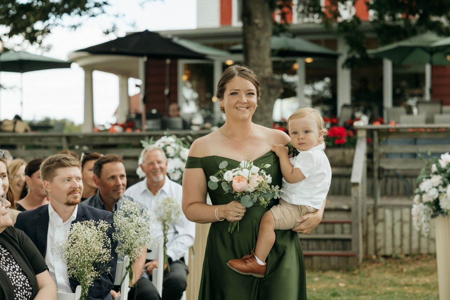 Bridesmaid in green off-shoulder dress carrying a baby and pink bouquet down the wedding aisle.