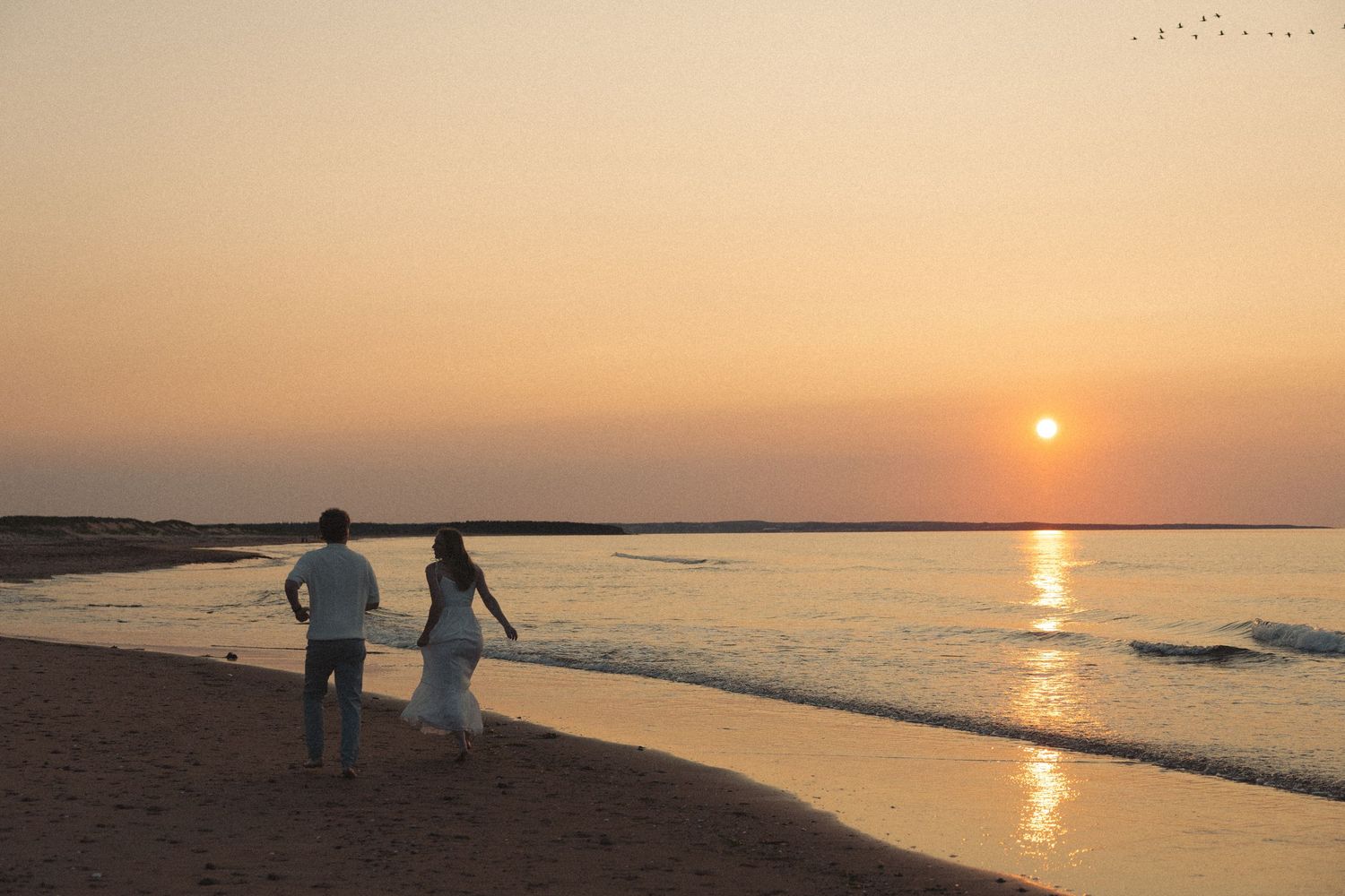 Romantic couple silhouetted walking along shoreline during vibrant golden sunset on beach.
