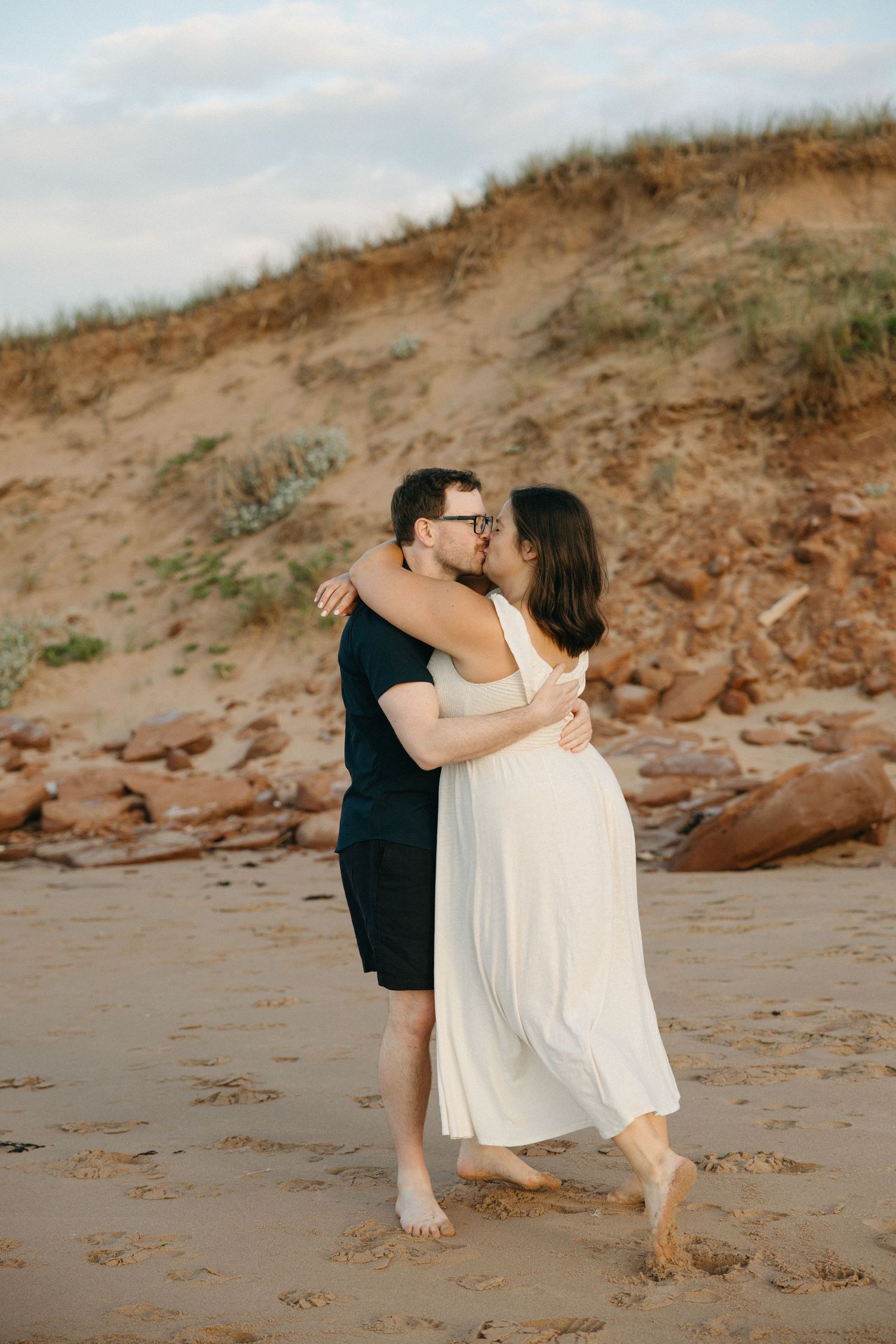 Couple kissing and embracing on sandy beach with golden hour light and dunes behind.