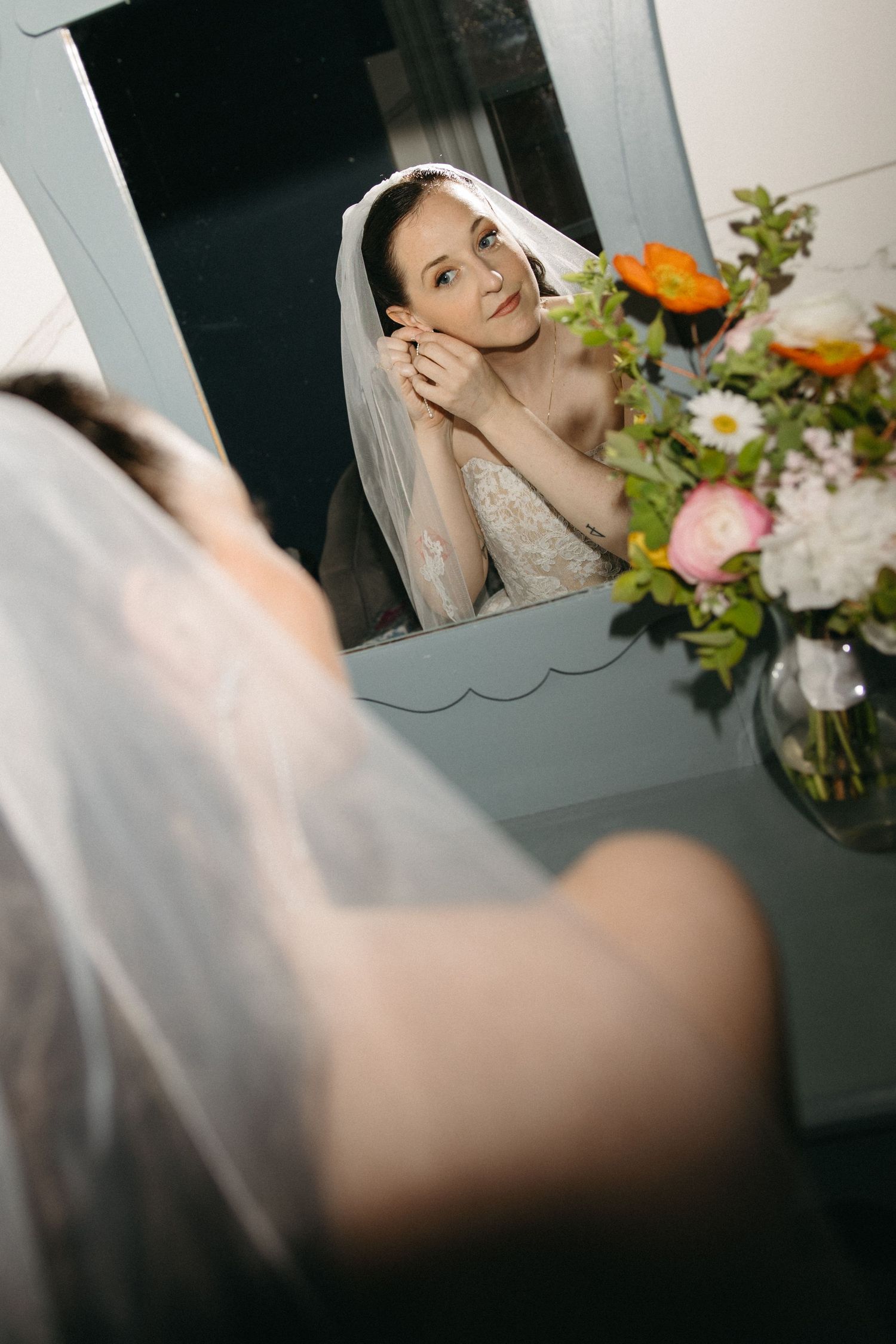 Bride in lace gown and veil adjusts earring while gazing into mirror beside fresh floral arrangement.