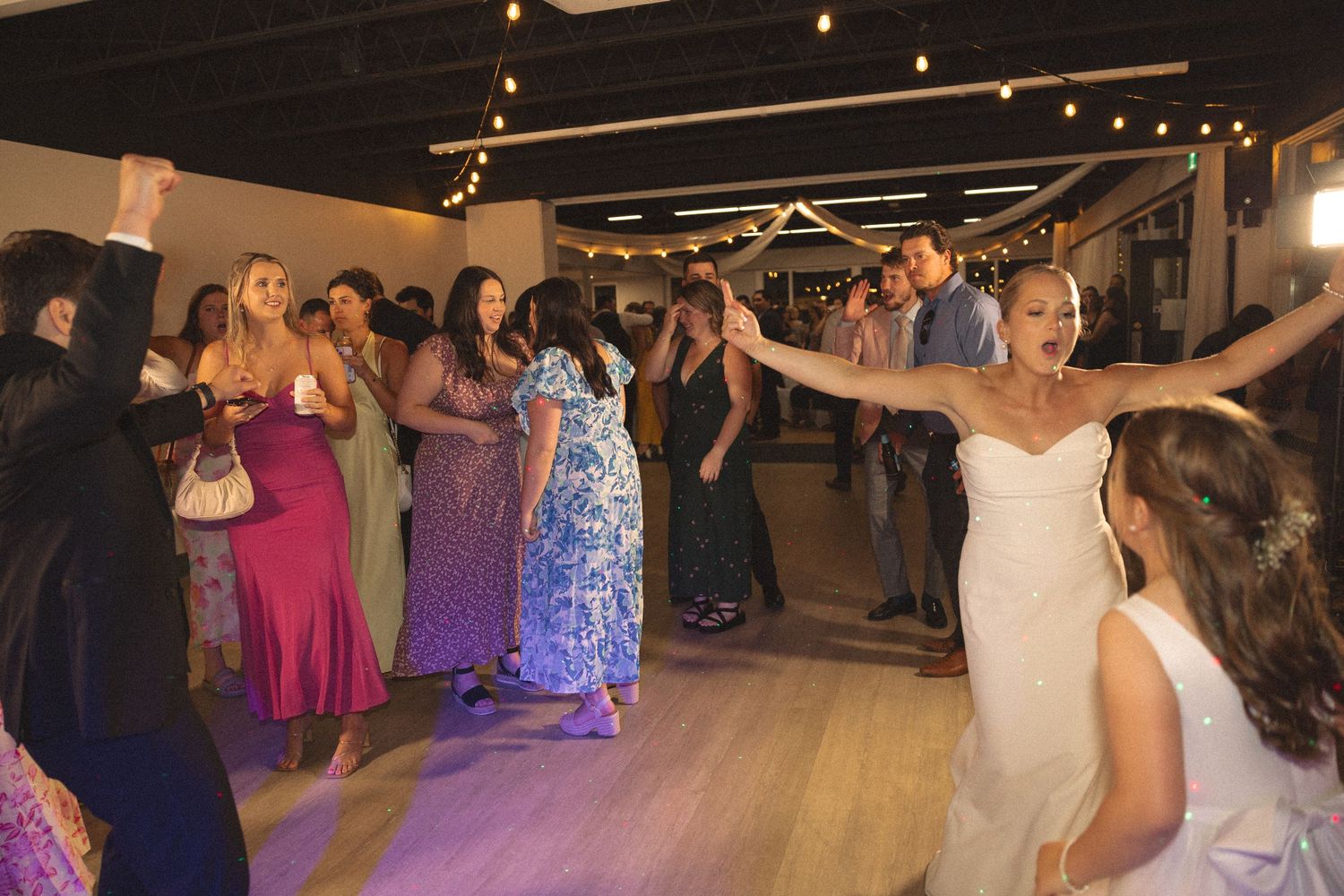 Wedding guests dance together on a lively dance floor under string lights at an indoor reception venue.