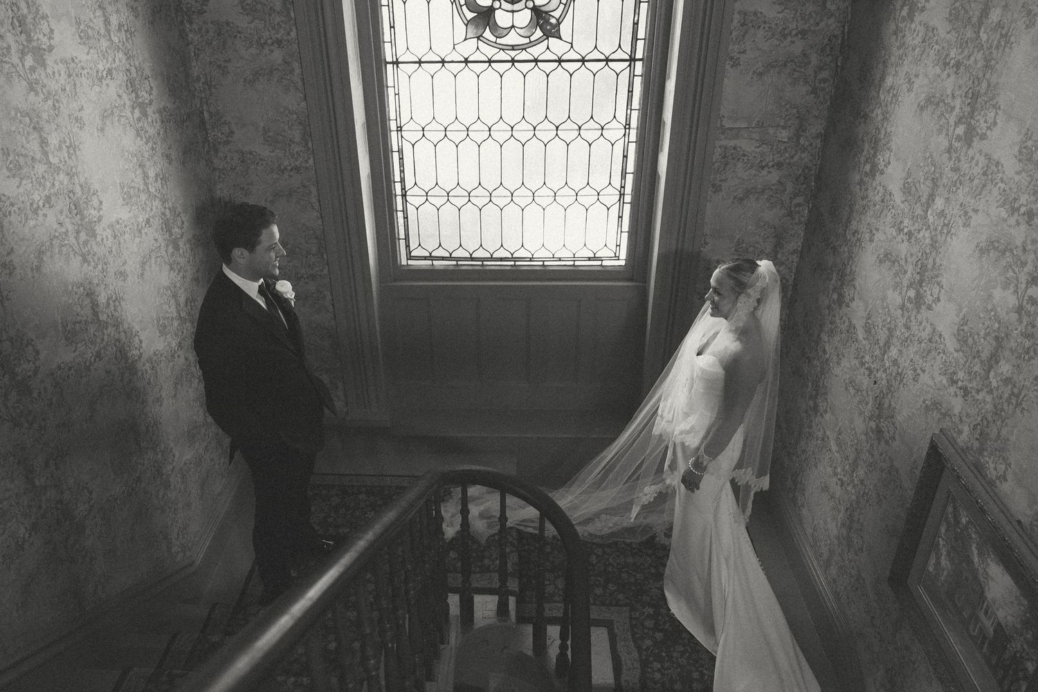 Black and white wedding photo on vintage staircase with dramatic window lighting.