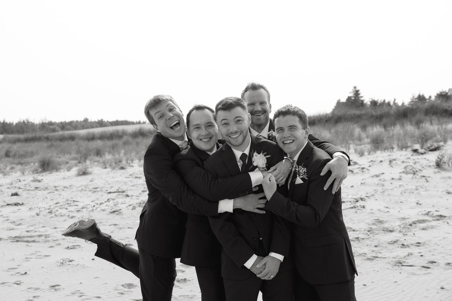 Groomsmen laughing and posing together on beach in formal attire.