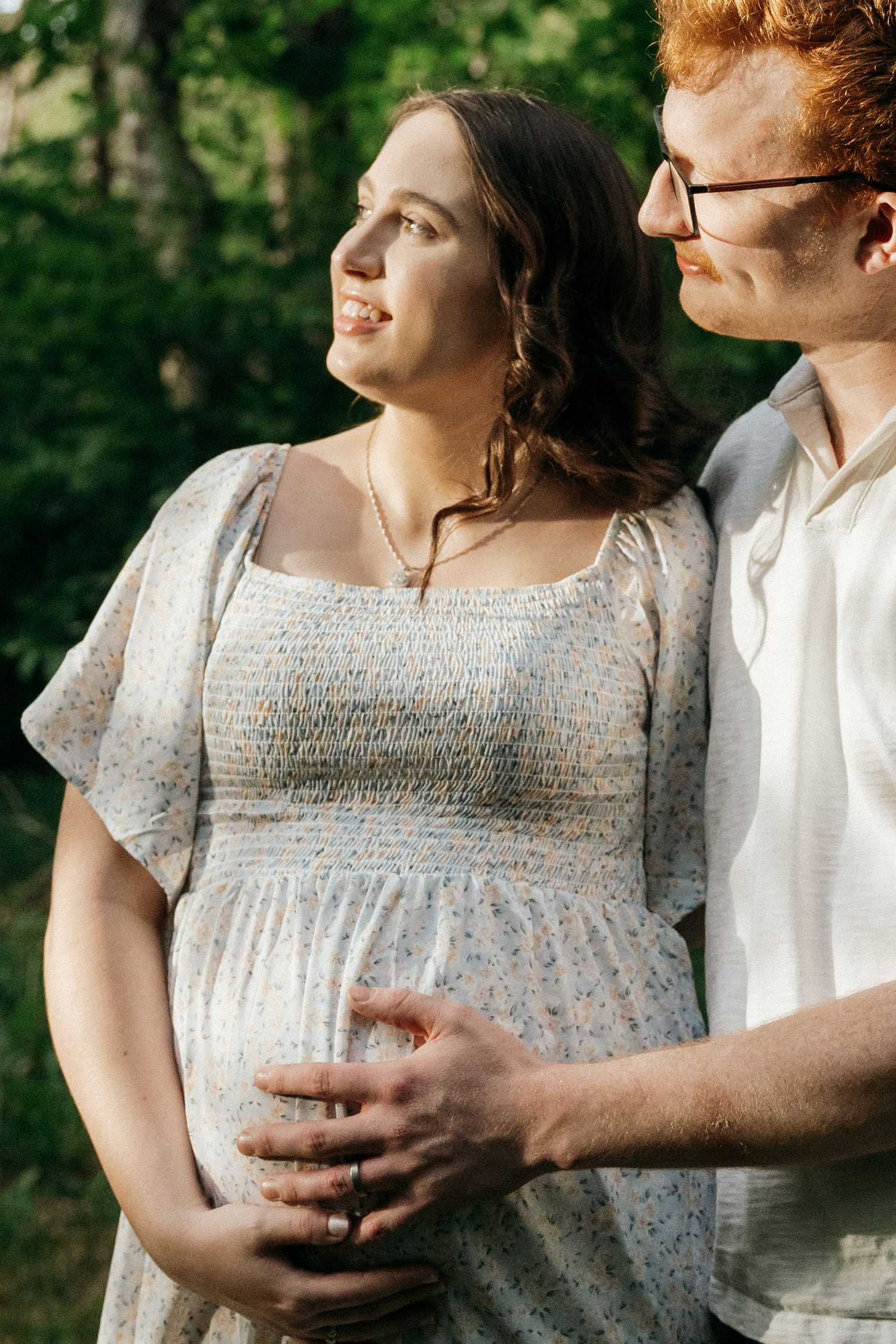 A tender maternity moment captured in a natural outdoor setting with soft lighting and greenery.