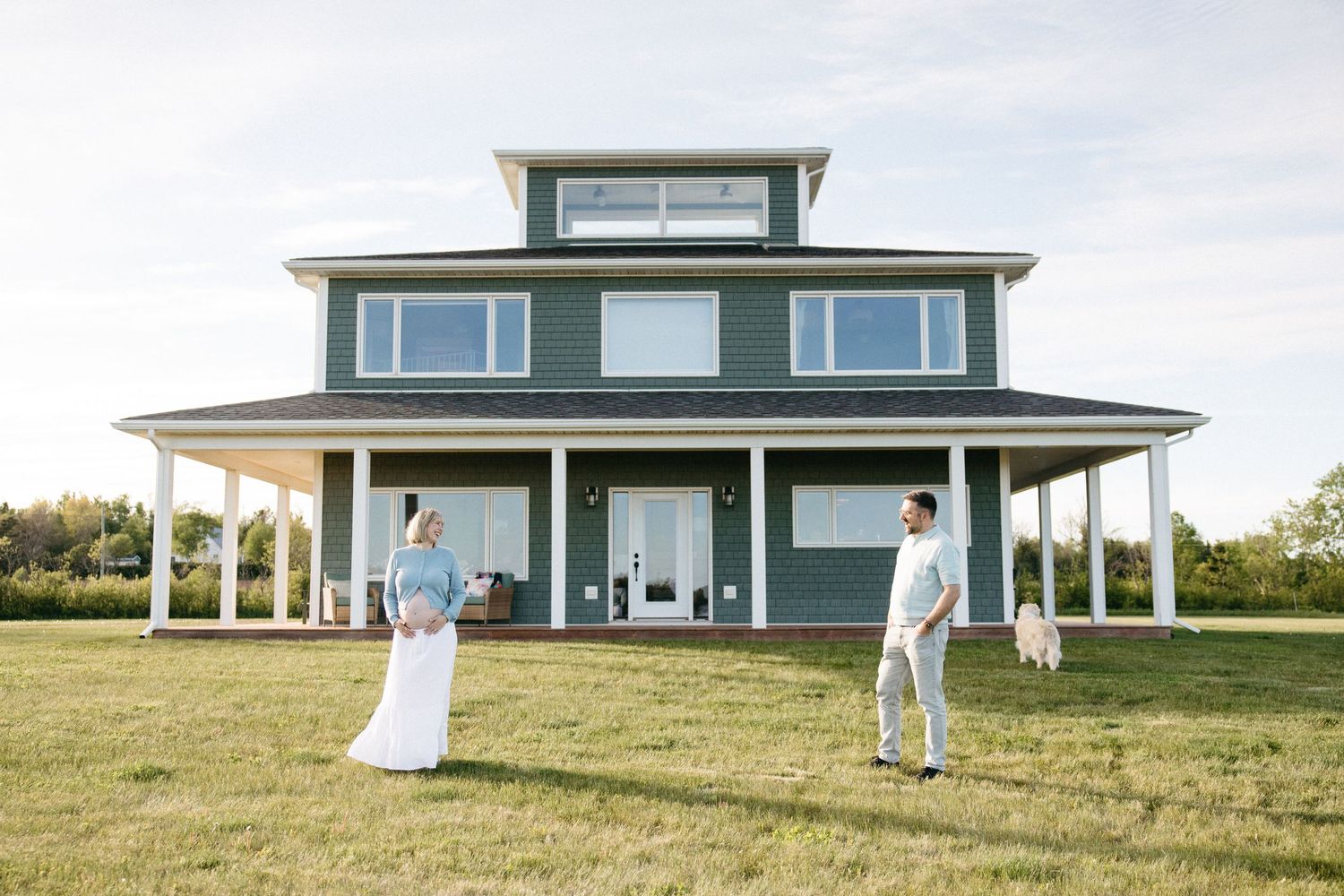 Couple standing in front of a large blue farmhouse with wraparound porch on a sunny day.