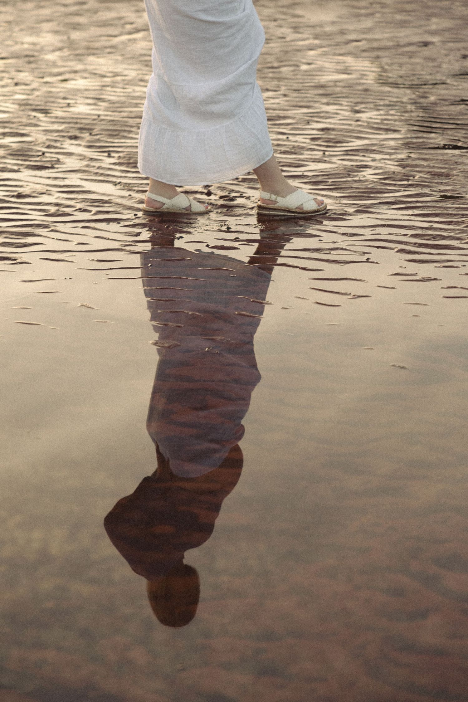 Person in white dress and sandals walking on wet reflective beach sand at golden hour.