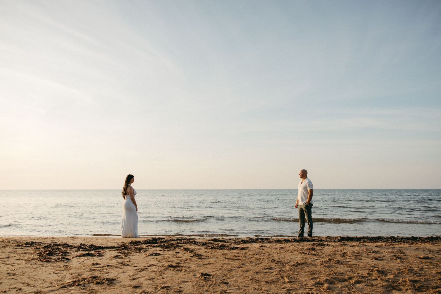 A couple stands apart on a sandy beach at sunset with calm ocean waves in the background.