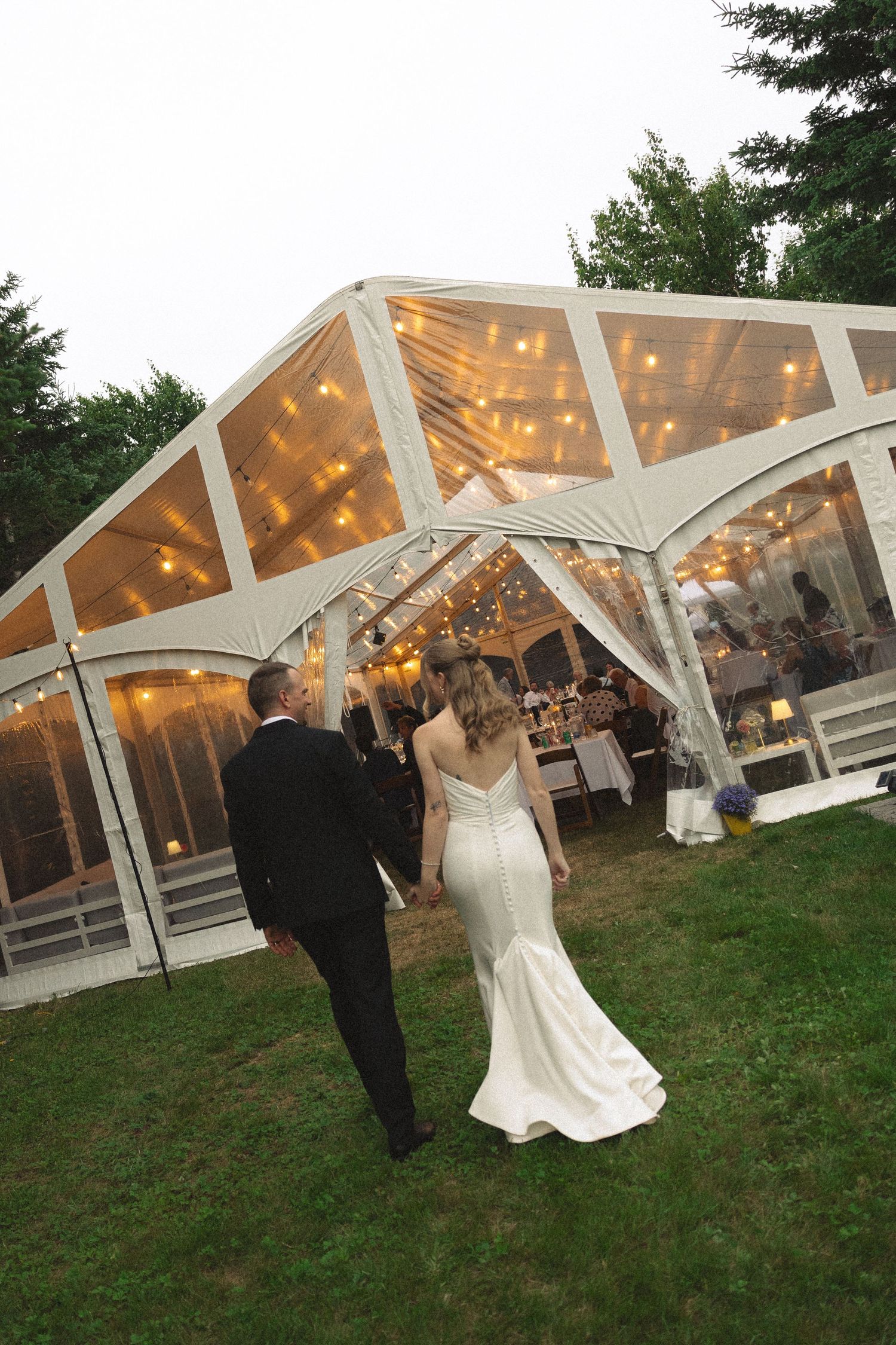 Bride and groom walking hand in hand toward a glowing transparent wedding tent adorned with string lights at dusk.