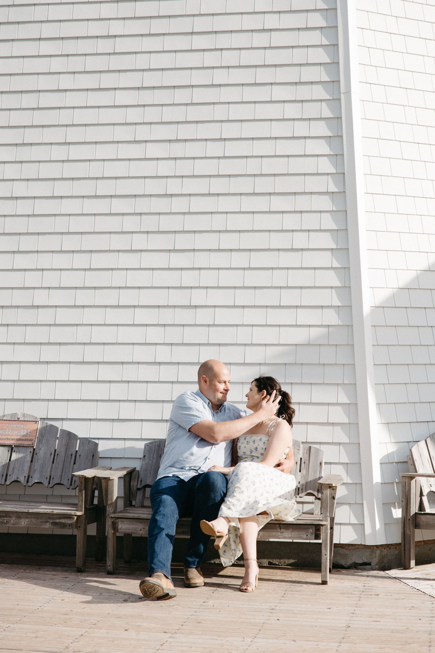 Romantic couple sharing tender moment seated on rustic wooden bench against white shingle wall.