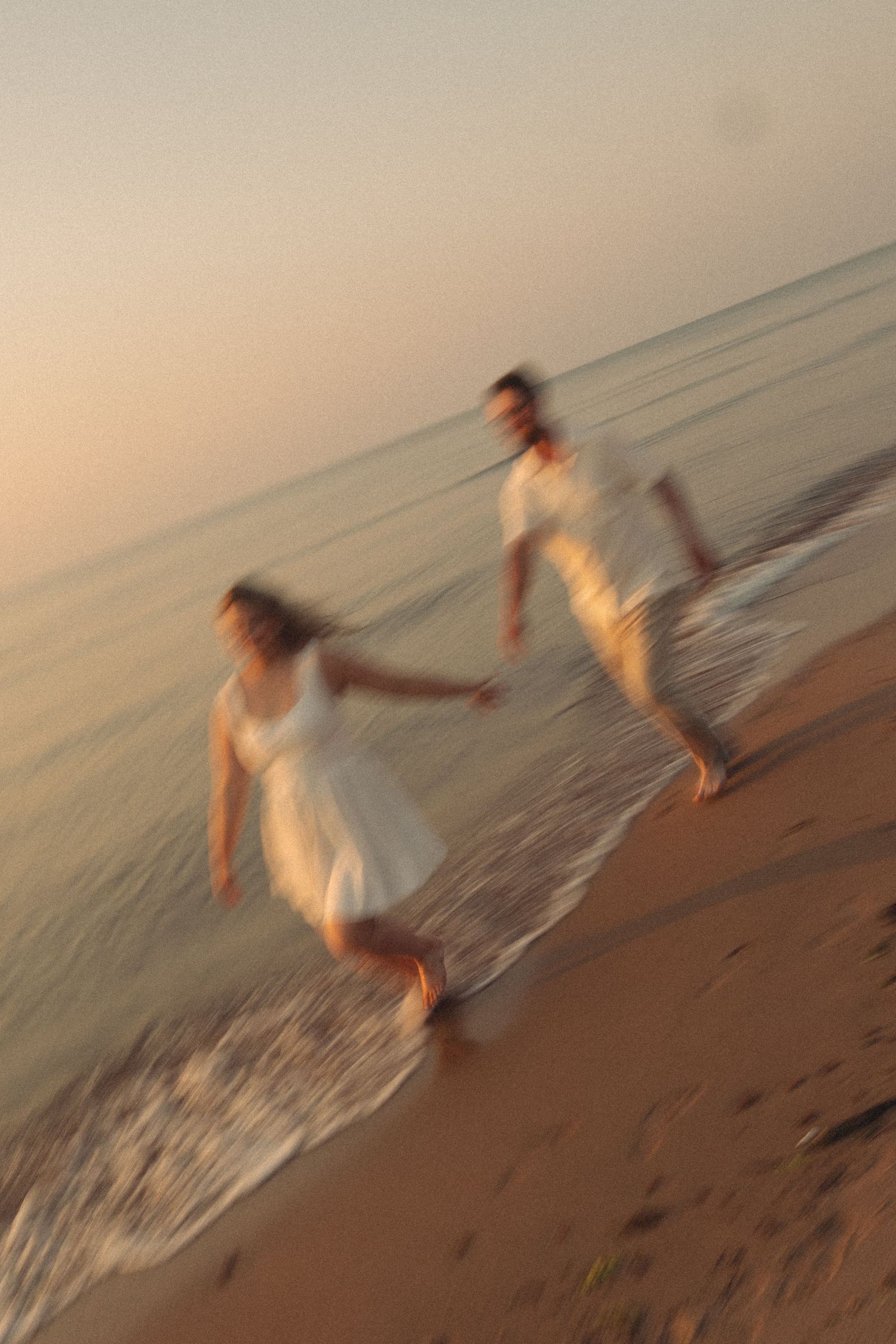 Couple in white outfits running hand-in-hand along a sandy beach shore at sunset.