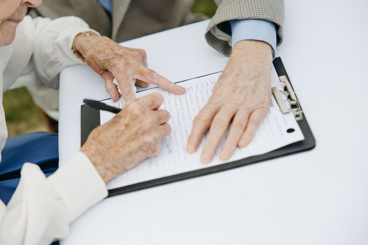 Two elderly hands review and sign a document on a clipboard at an outdoor wedding ceremony table.