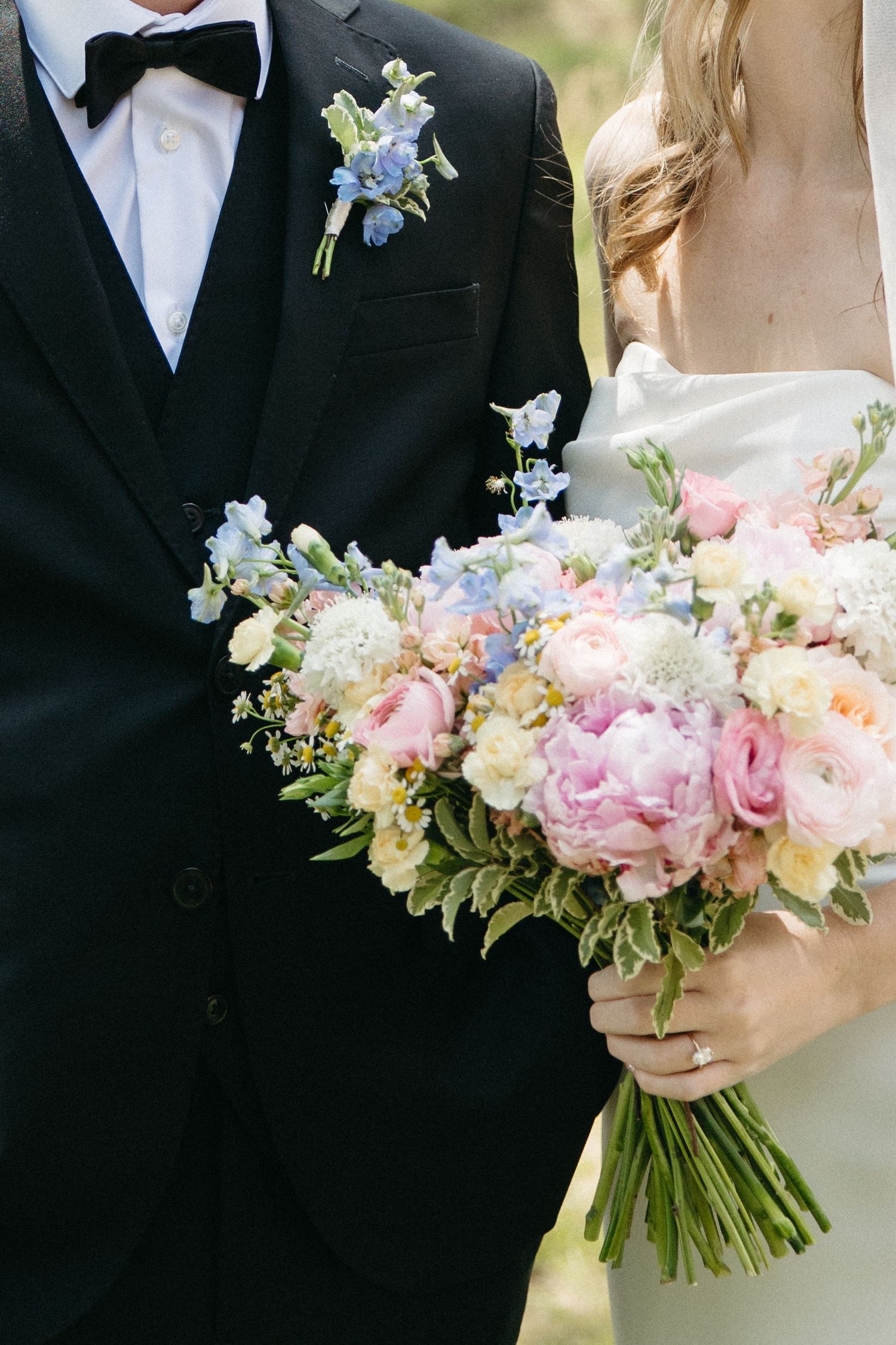 Bride holding a lush pastel wedding bouquet with pink peonies and blue flowers beside groom in black tuxedo.