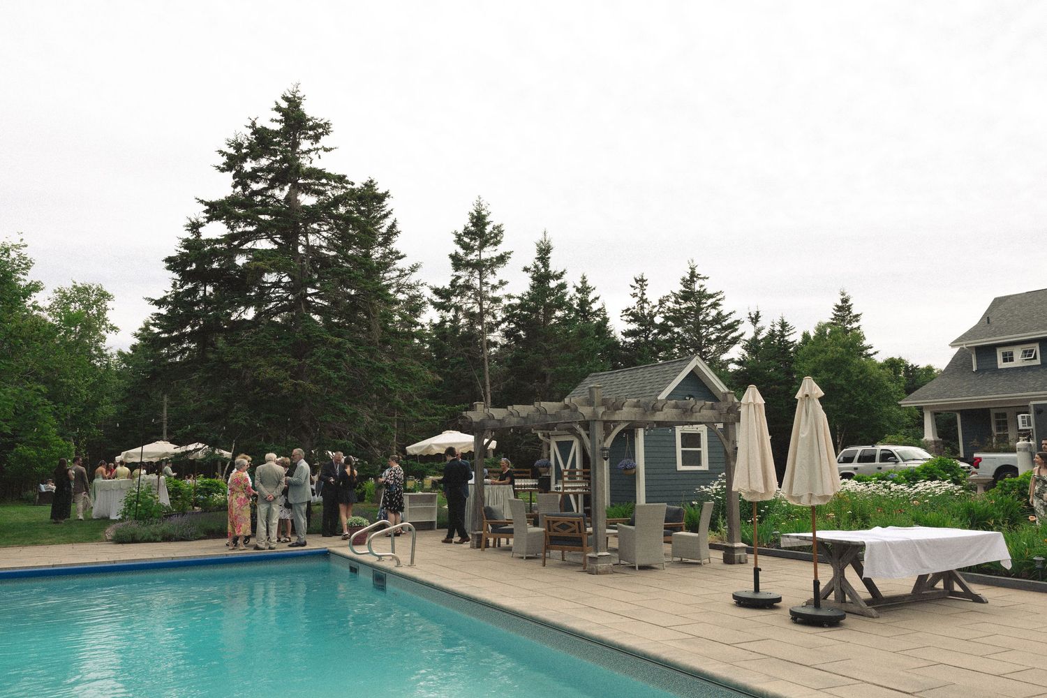Wedding guests mingle poolside at elegant outdoor reception with white umbrellas and lush green trees.