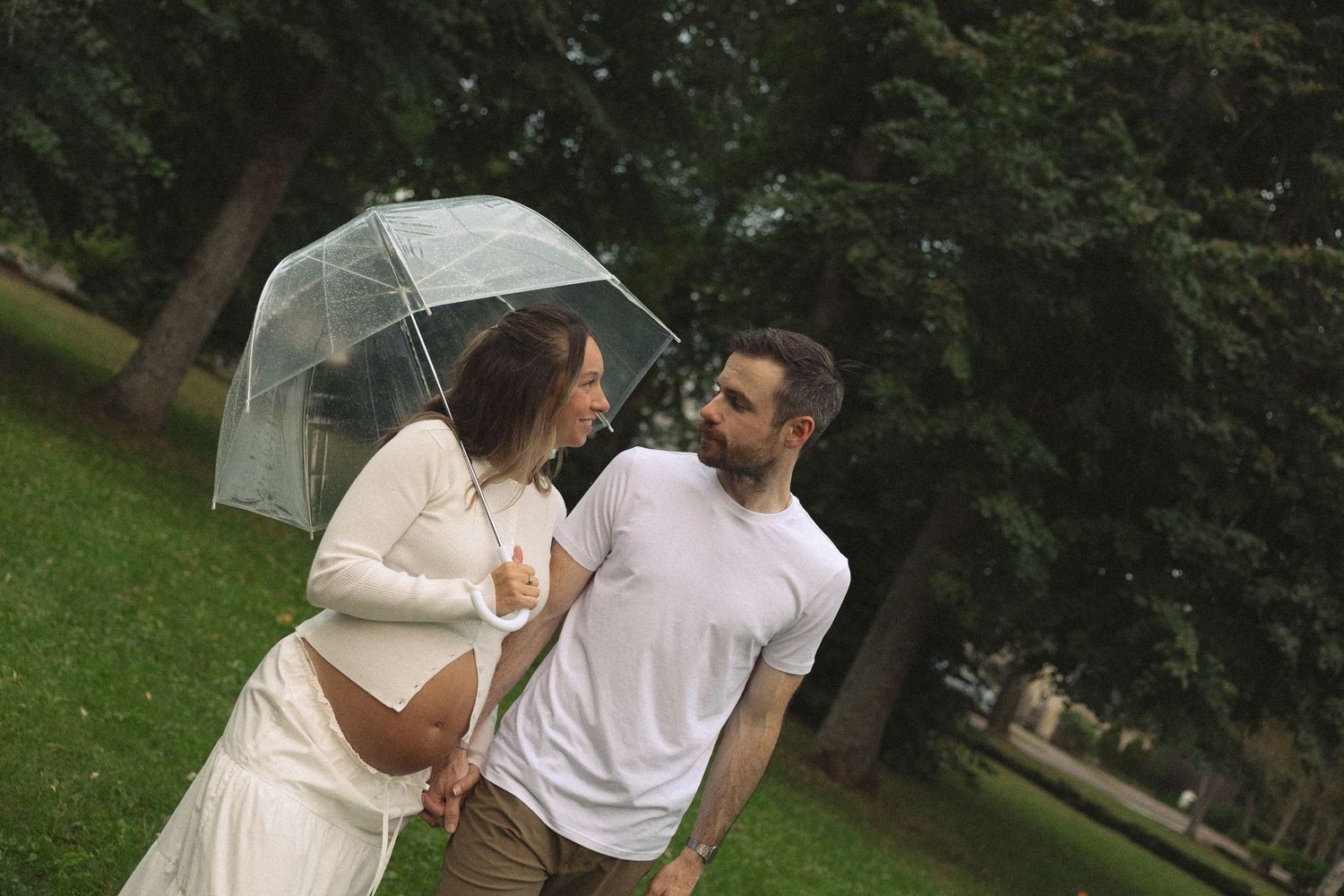 Couple sharing umbrella in park, pregnant woman in white dress with bearded man.
