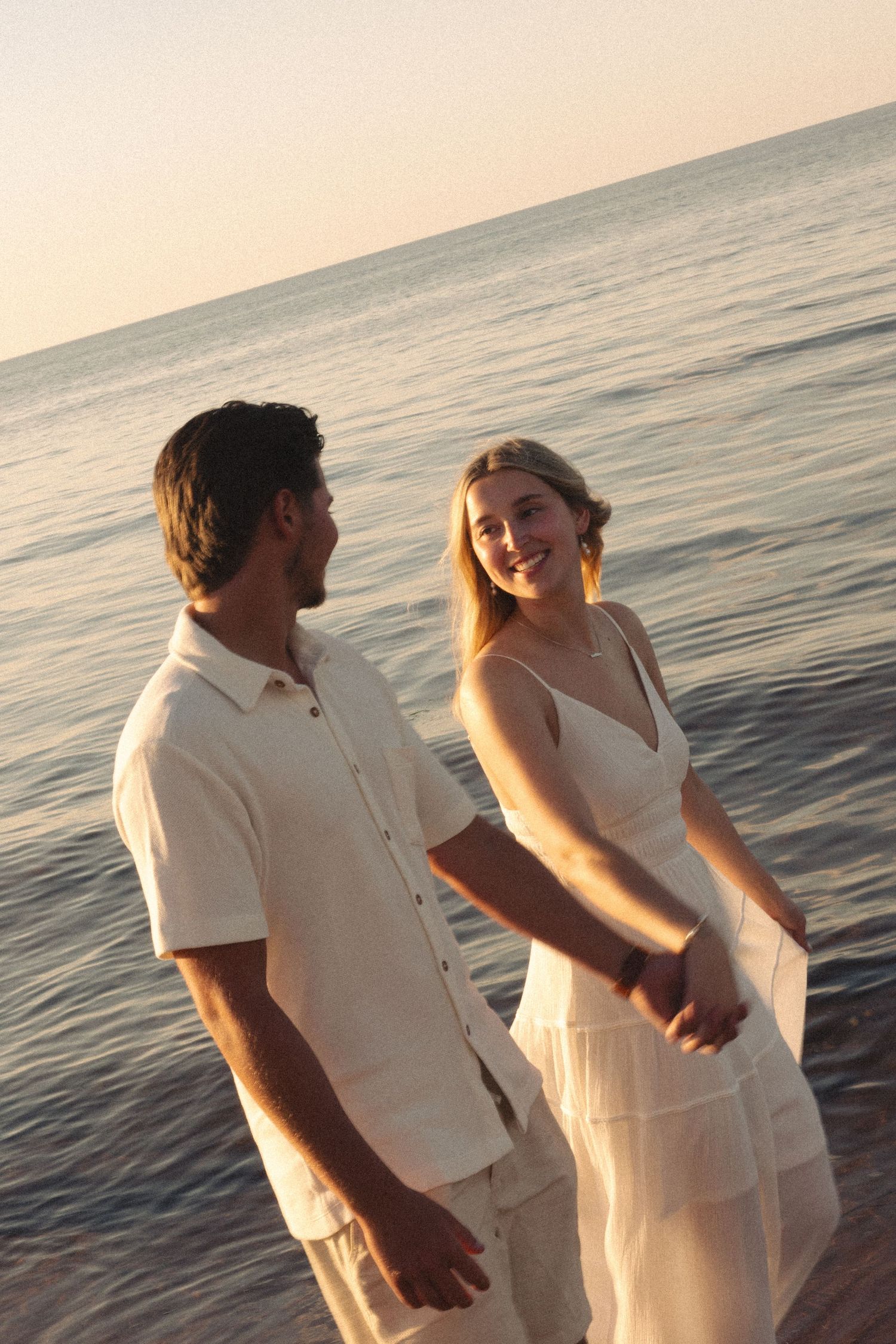 A smiling couple in matching white outfits hold hands on a beach at sunset with calm ocean waters behind them.