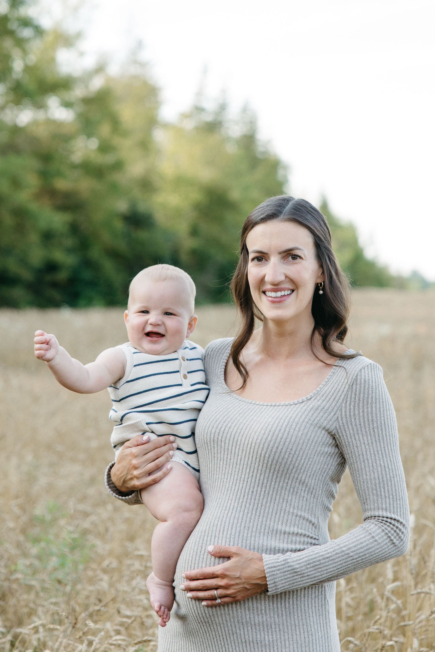 Smiling pregnant woman in gray dress holds laughing baby in striped onesie in golden wheat field.