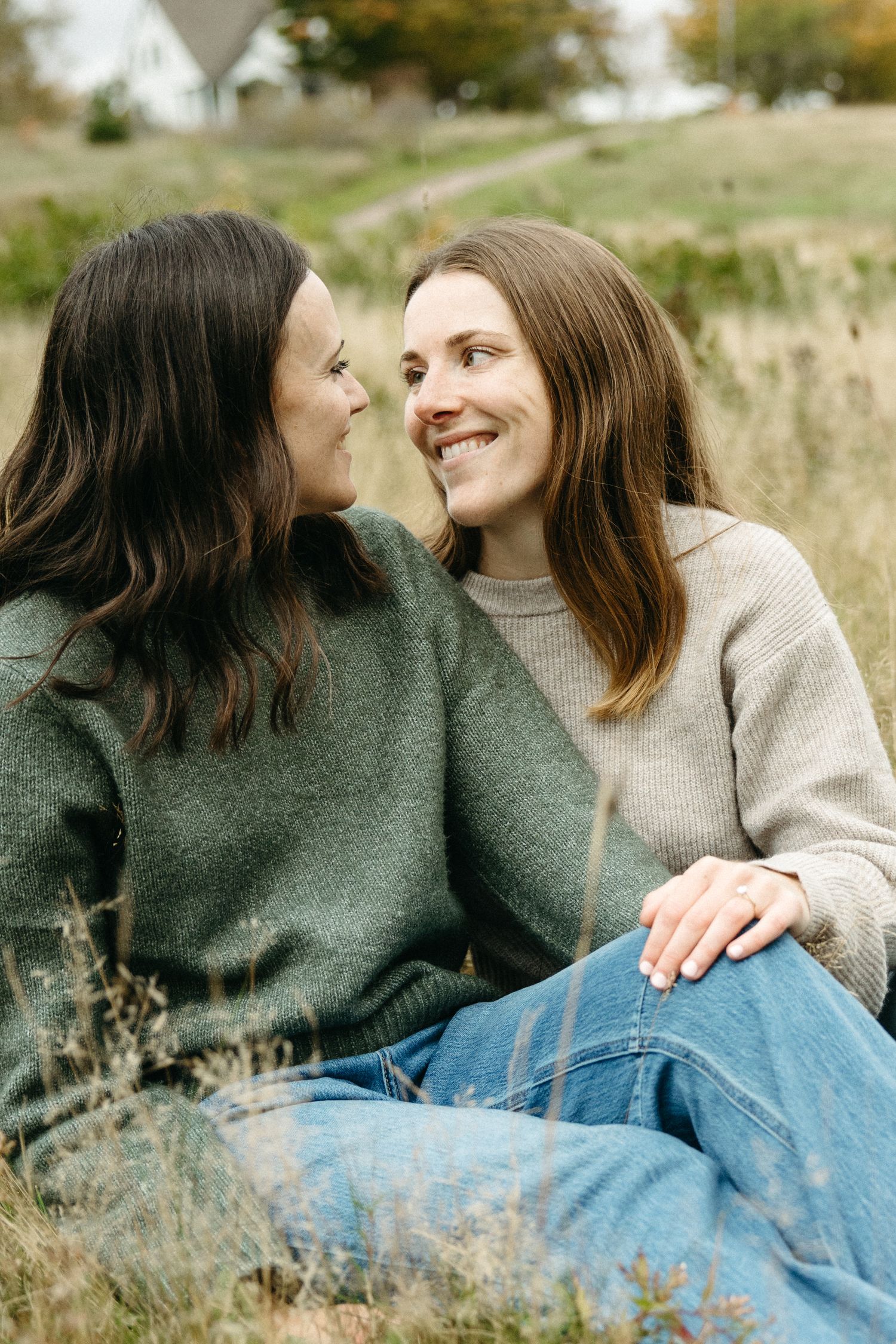 Two women with long hair smile at each other while sitting close together in a grassy autumn field.