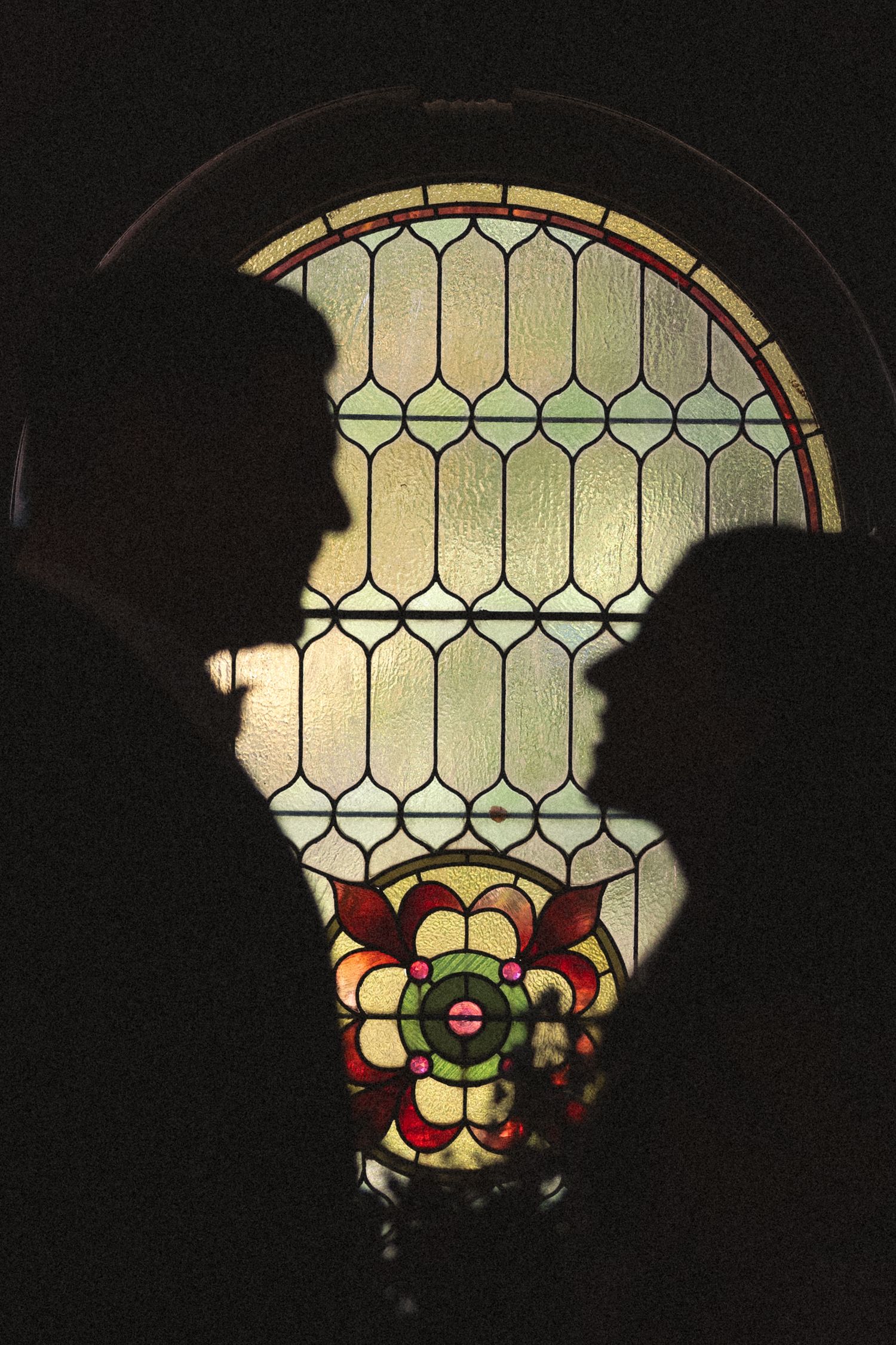 Silhouettes of two people face each other in profile before an arched stained glass window with a colorful red and green floral medallion.