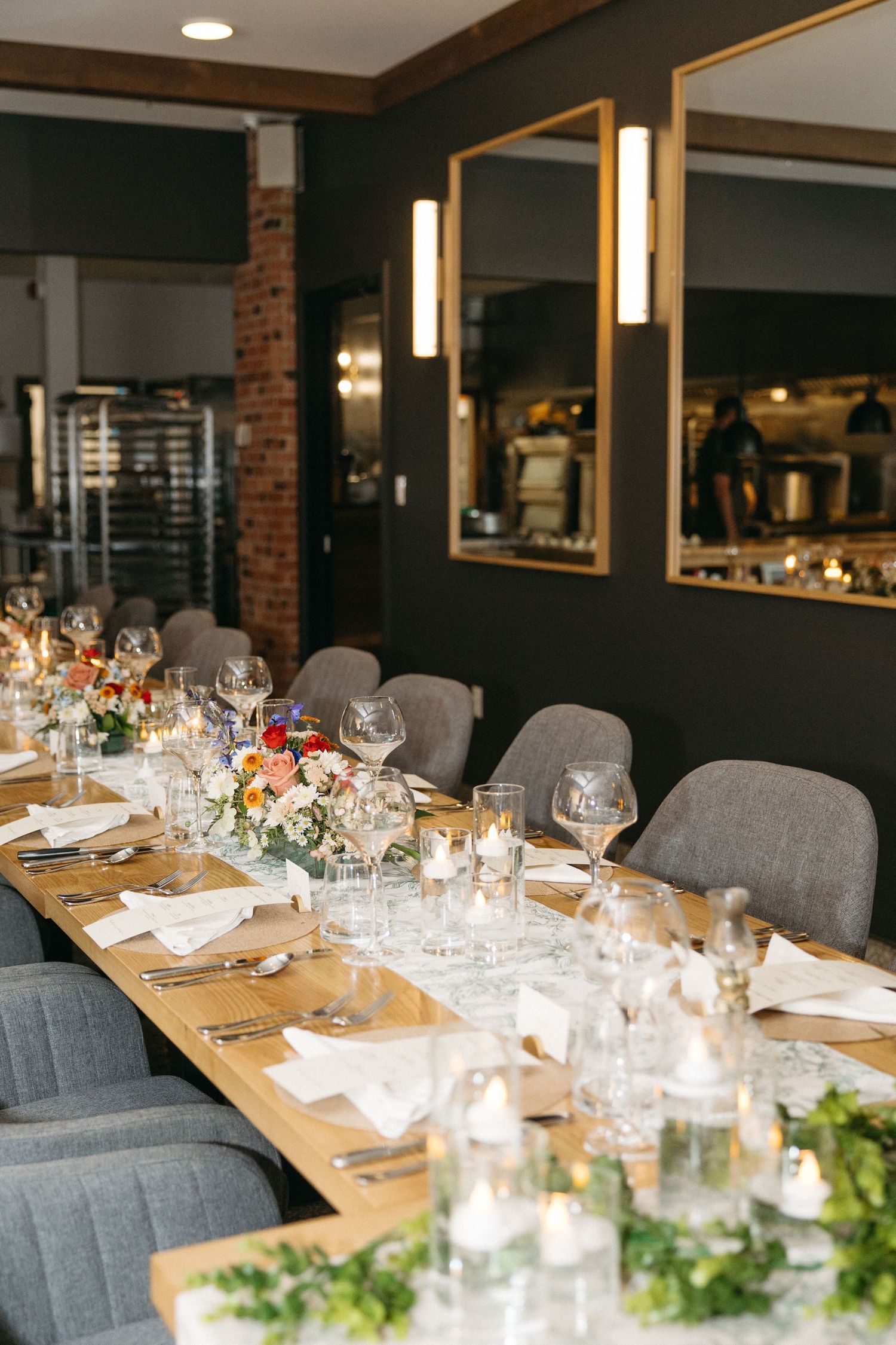 Long wooden banquet table set with floral centerpieces, candles, wine glasses and gray chairs in a modern restaurant with brick walls.