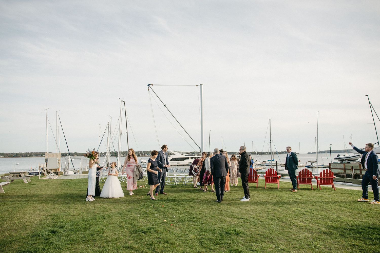 Wedding guests gather on a green lawn beside a marina with sailboats and red chairs under a soft, cloudy sky.