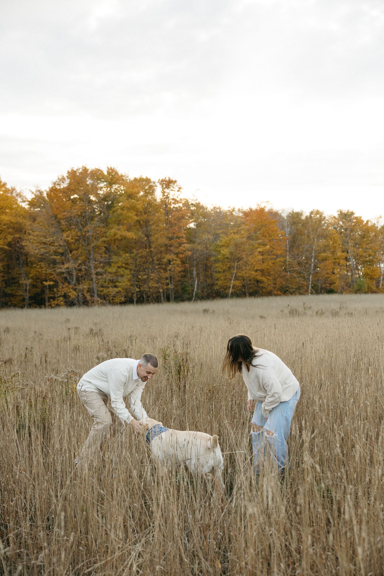 Couple playing with their white dog in tall grass field during autumn.