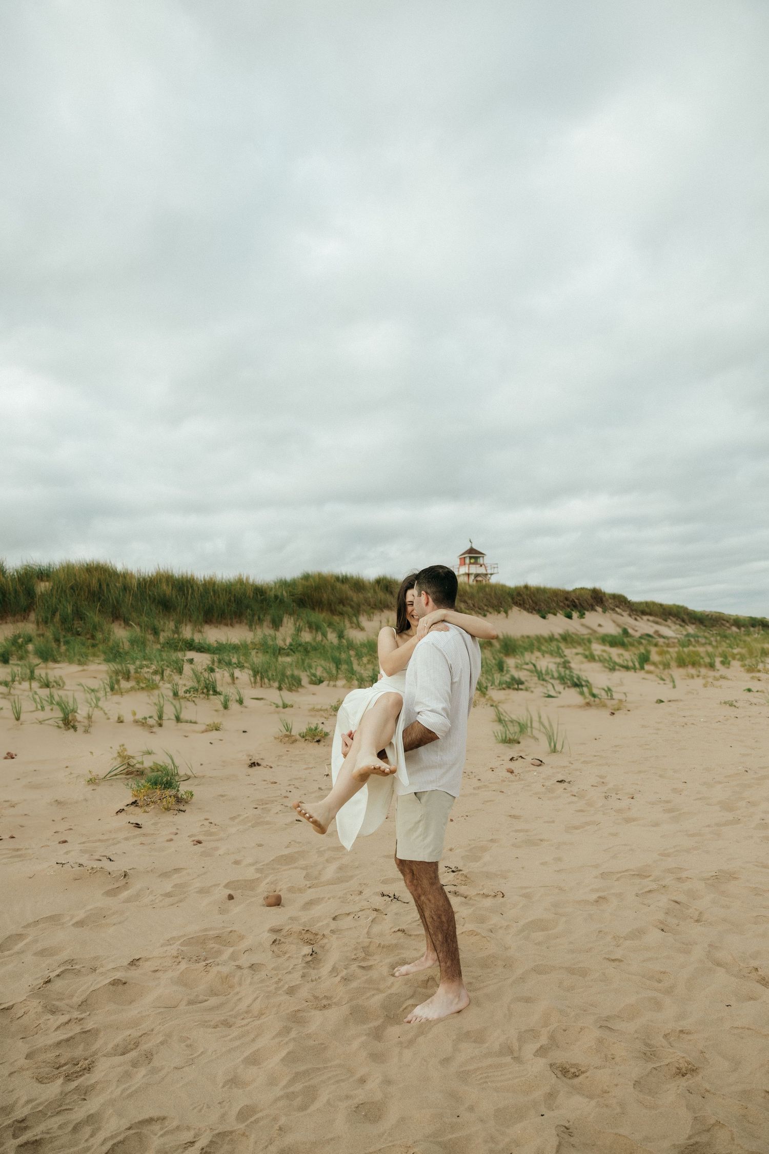 A couple playfully poses on a beach with sand dunes and cloudy skies in the background.