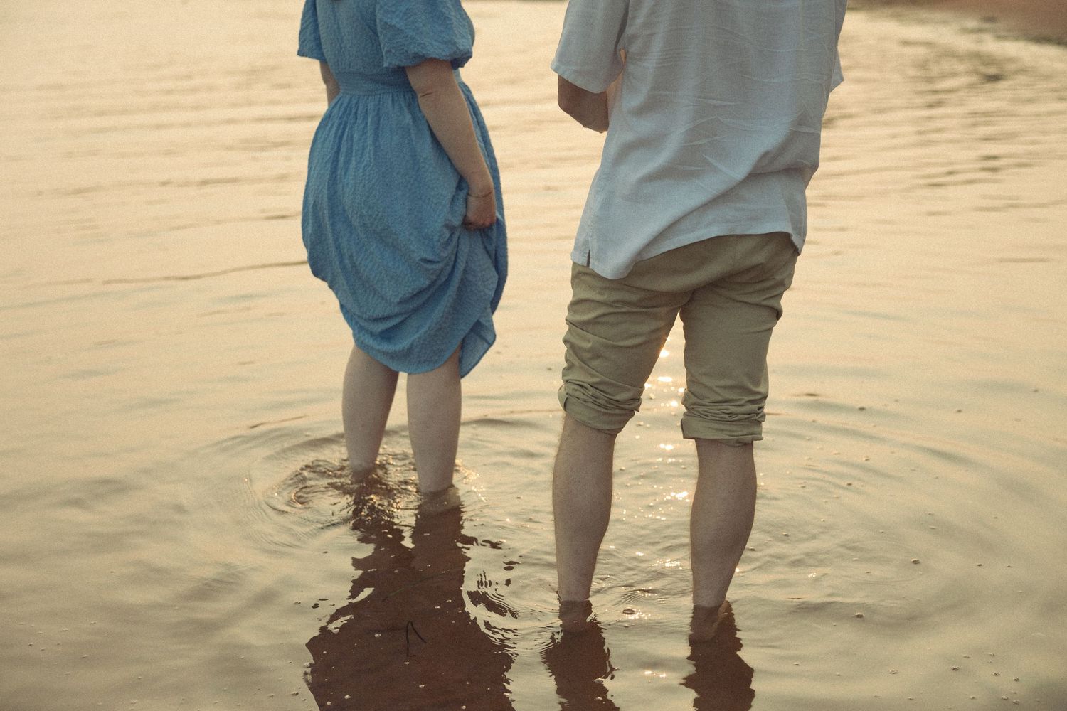 Couple standing ankle-deep in ocean water during sunset, wearing casual blue and white clothing.